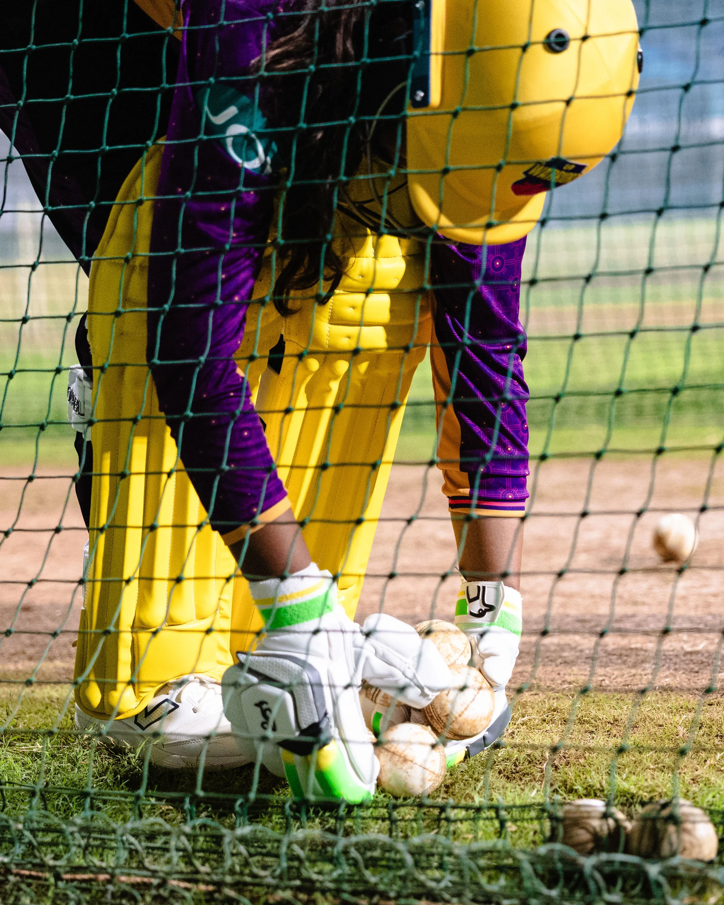 Close-up of a soccer goalkeeper in yellow and purple goalkeeper gear, kneeling inside the goal with multiple soccer balls at their feet, behind a green goal net on a grassy field.