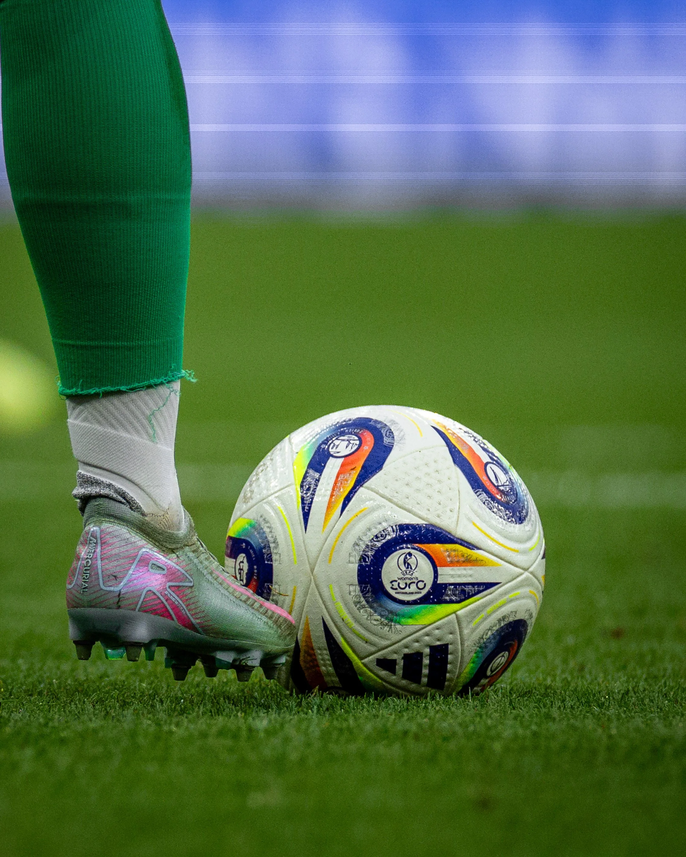 Close-up of a soccer player’s foot in a pink and white cleat pressing a colorful soccer ball on a green grass field.