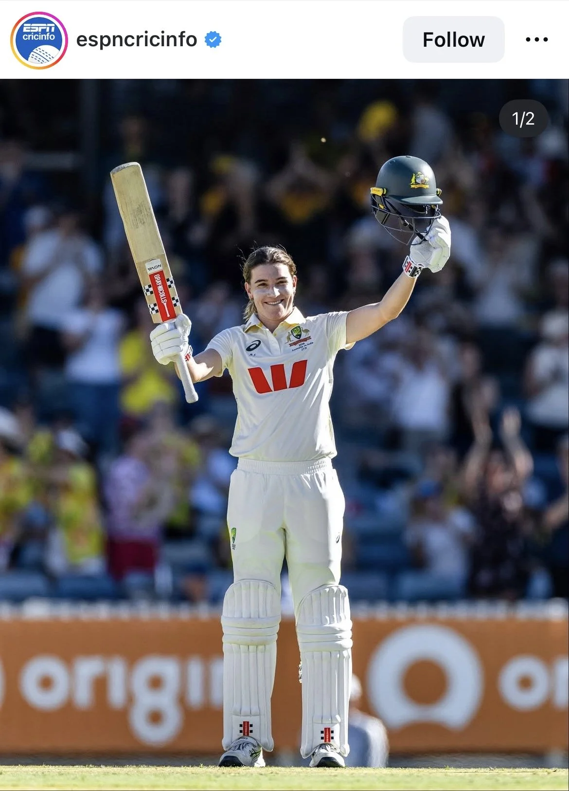 A female cricket player in white uniform holding a cricket bat and helmet, raising her helmet with a smile on her face during a cricket match.