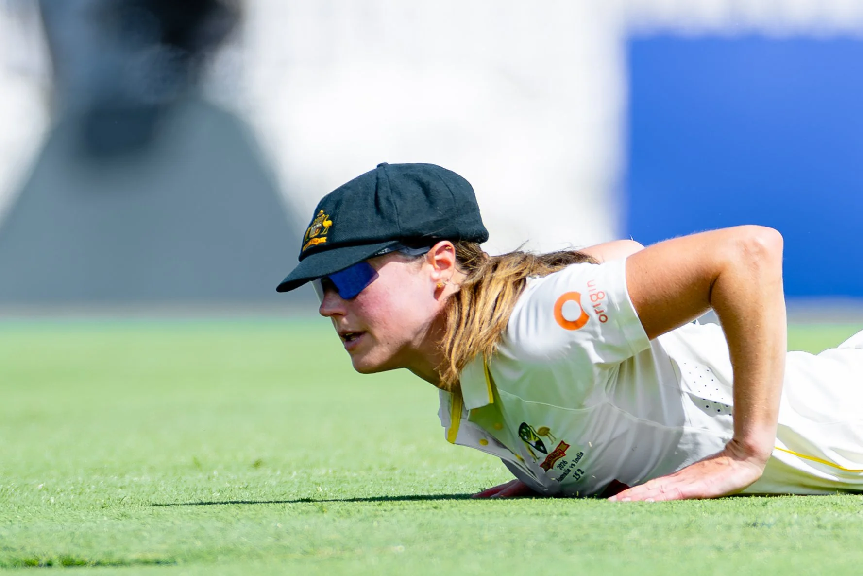 A female athlete in cricket attire doing a push-up on a grassy field, wearing a black cap, sunglasses, and a white sports uniform with logos.