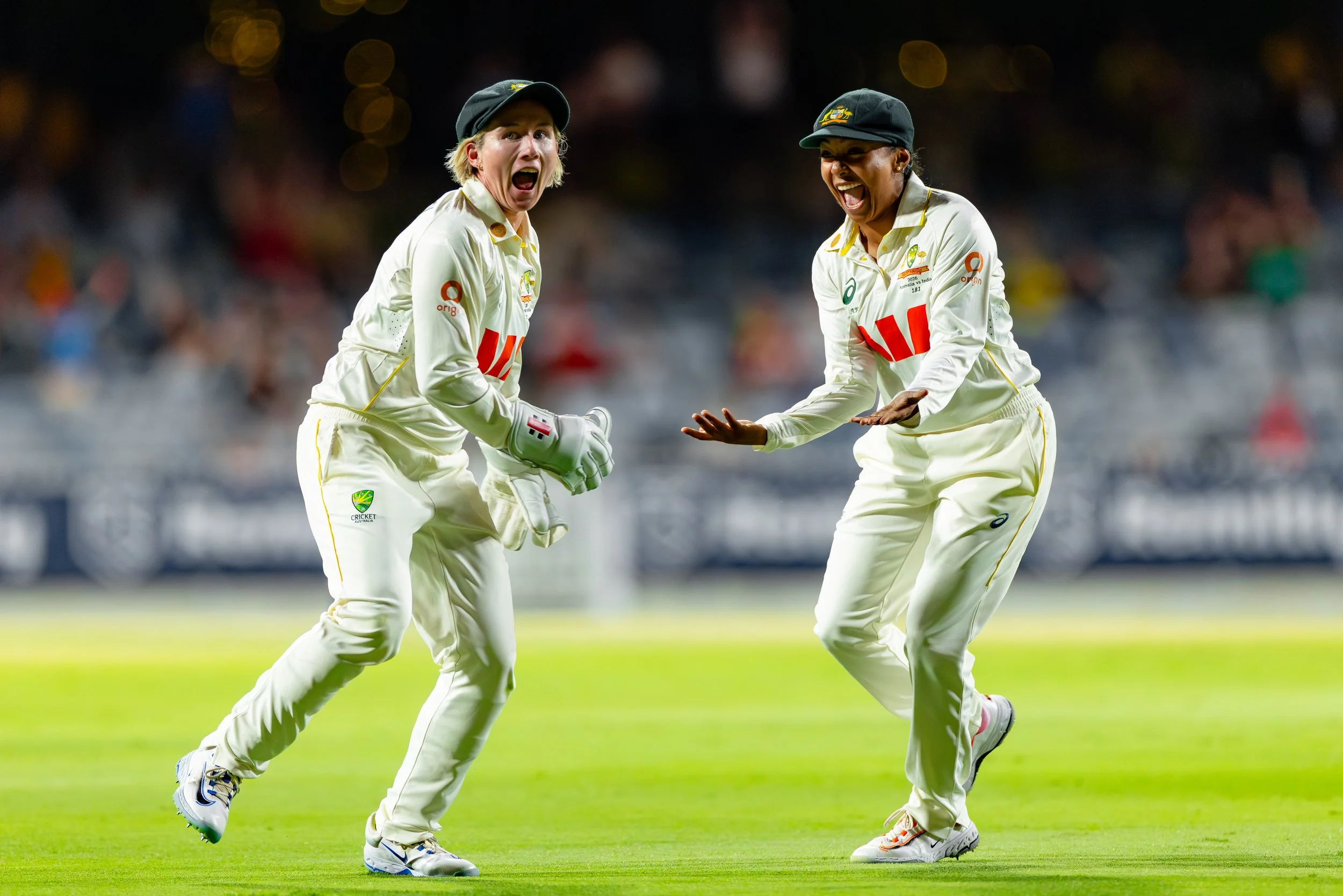 Two female cricket players celebrating on the field, wearing white uniforms with Australia team logos, with a blurred stadium crowd in the background.