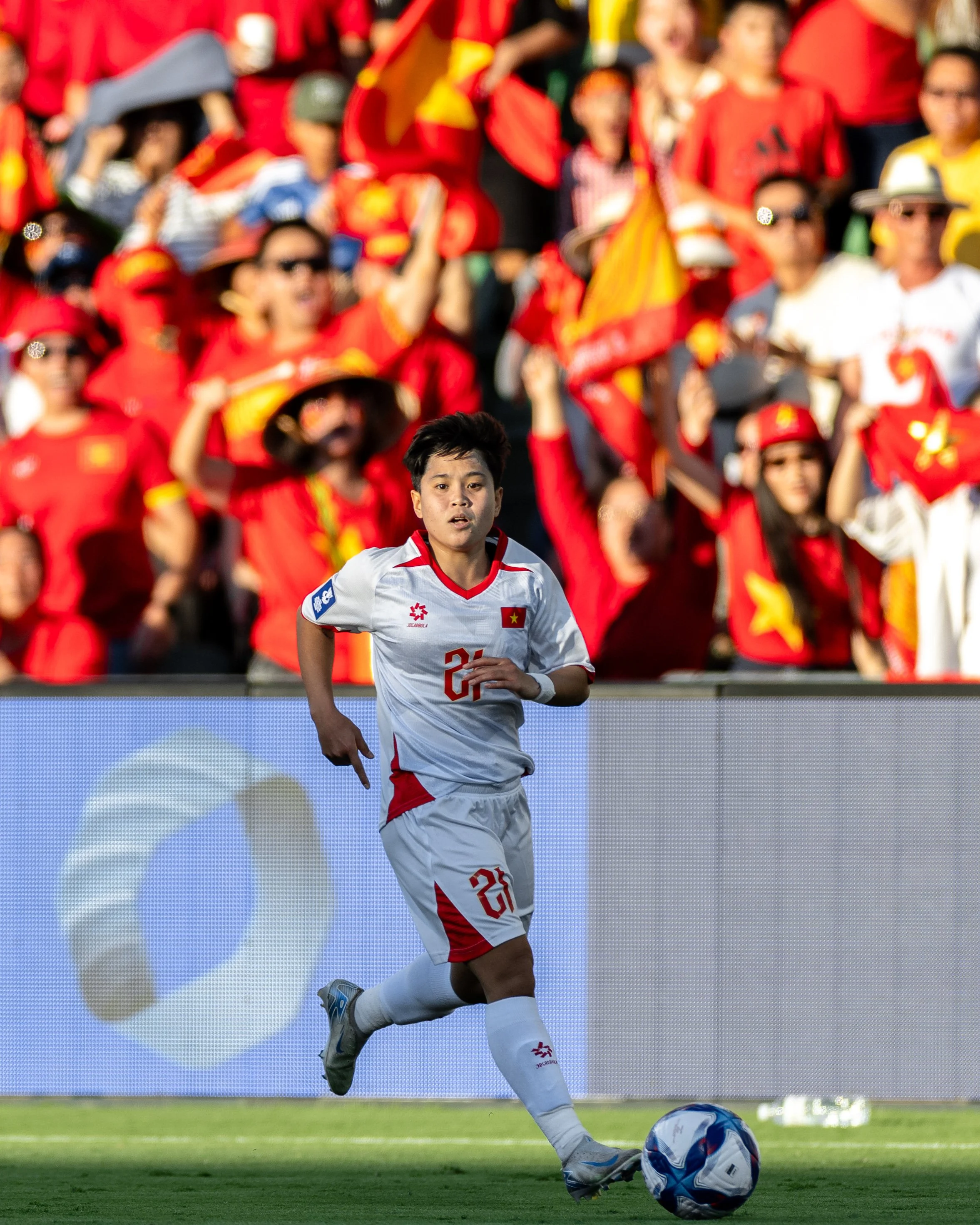 A soccer player wearing a white uniform with red details, number 23, and the Vietnam flag on the chest, runs on the field and kicks a soccer ball, with a crowd of spectators in red and yellow shirts cheering in the background.