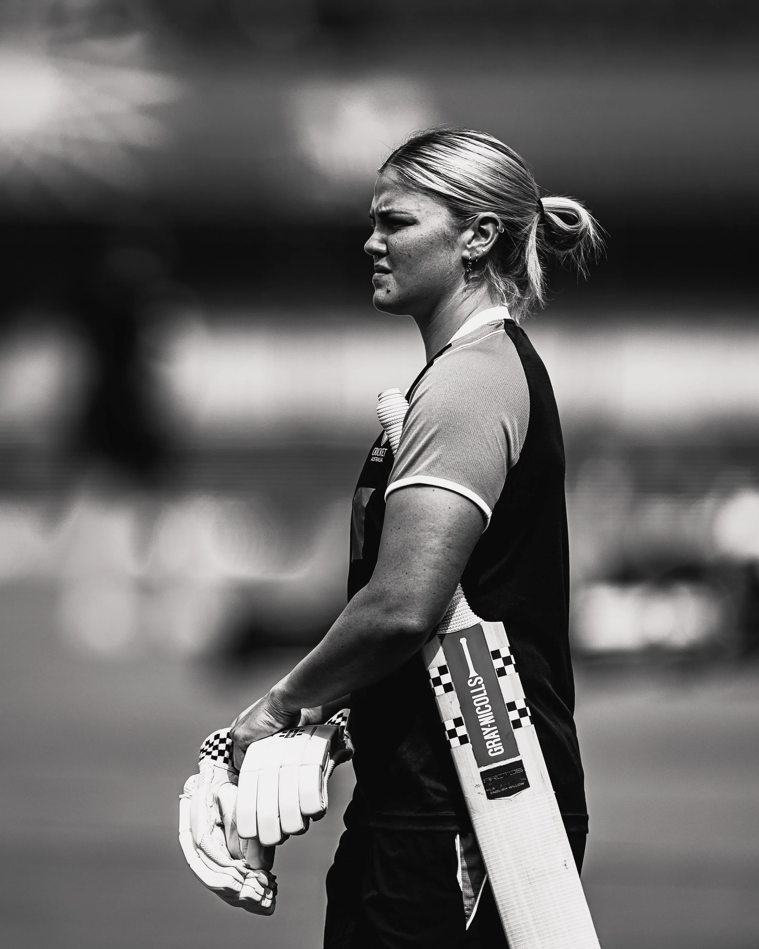 Black and white photo of a female cricket player wearing gloves and holding a cricket bat, standing outdoors with a cloudy sky in the background.