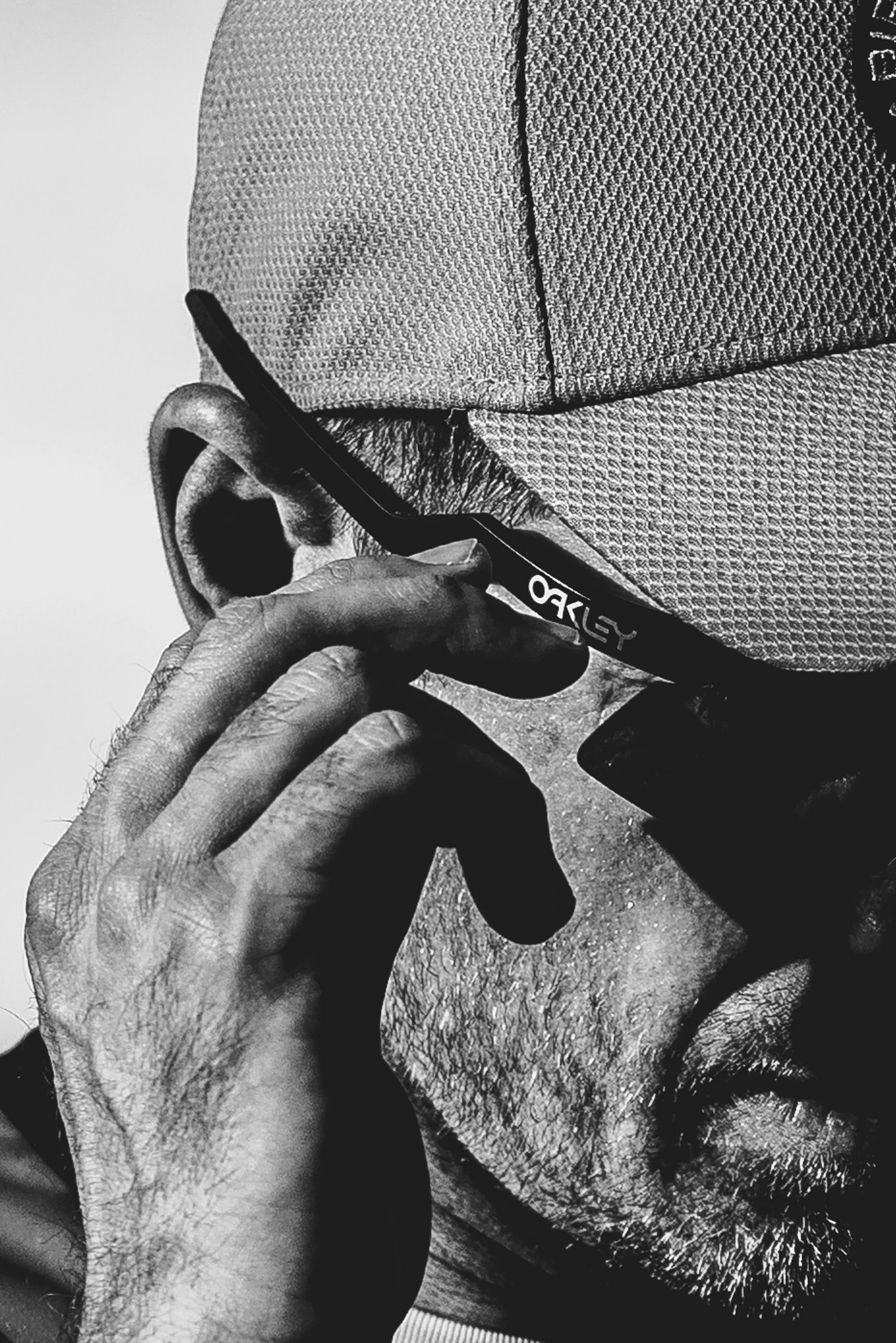 Close-up black and white photo of a person holding a smartphone near their face, with a prominent beard and textured clothing visible.