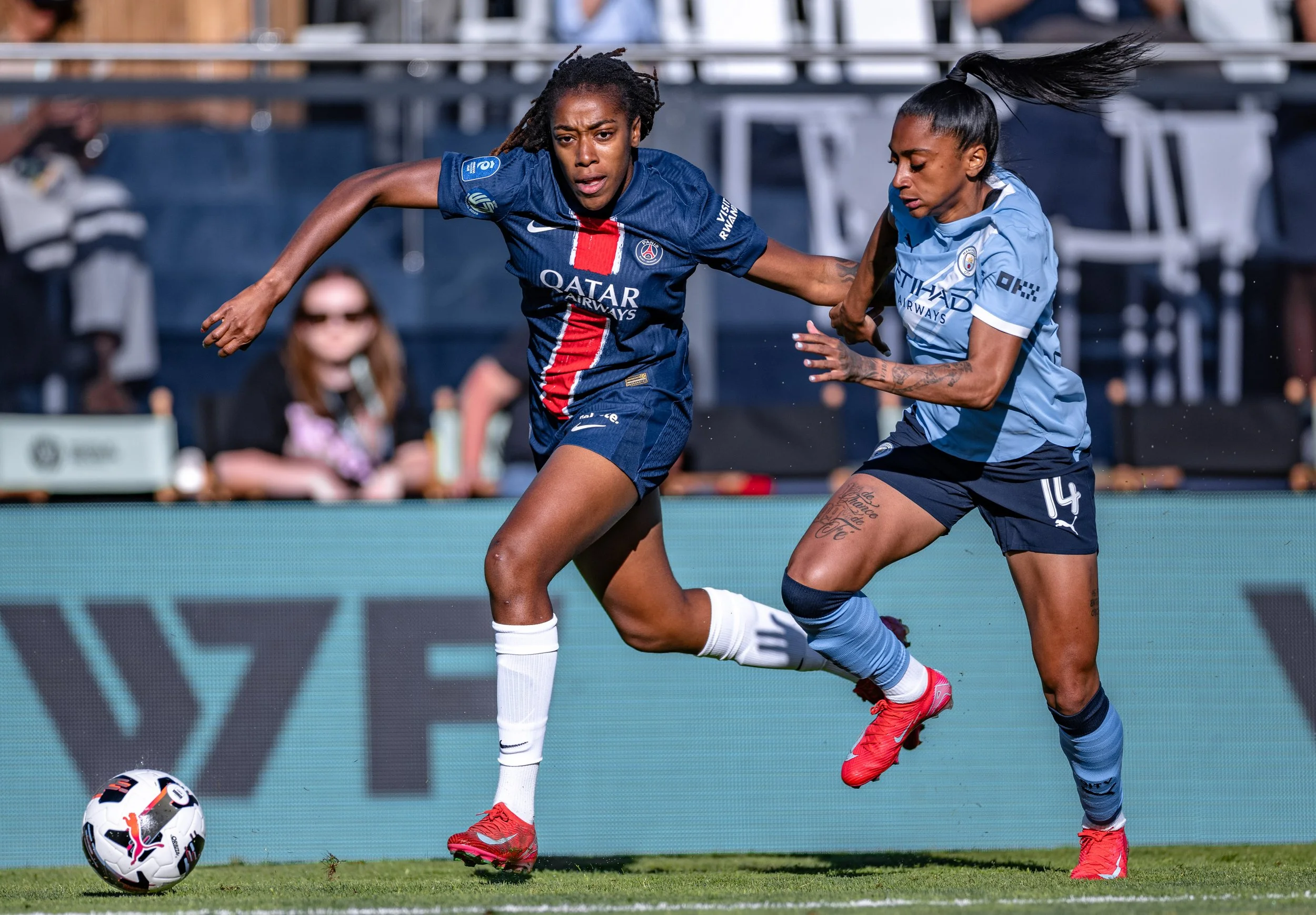 Two women playing soccer. The woman on the left is wearing a dark blue jersey with red and white stripes, and the woman on the right is in a light blue jersey. They are competing for ball possession on a green field, with a person in sunglasses sitti