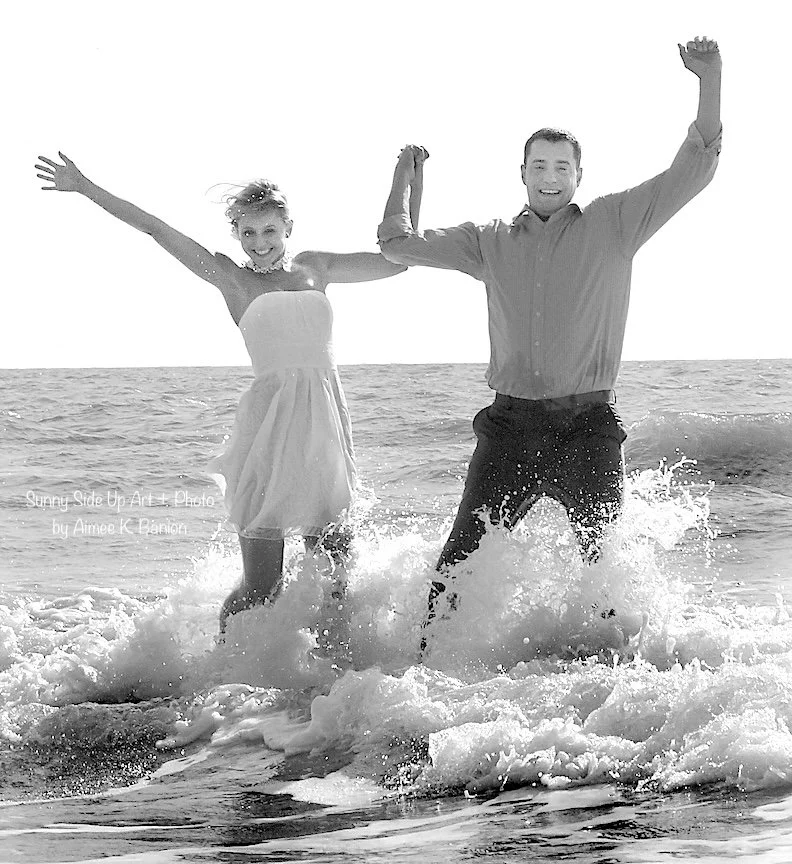 A bride and groom holding hands and jumping in the ocean waves, smiling, with their arms raised in a black and white photo.