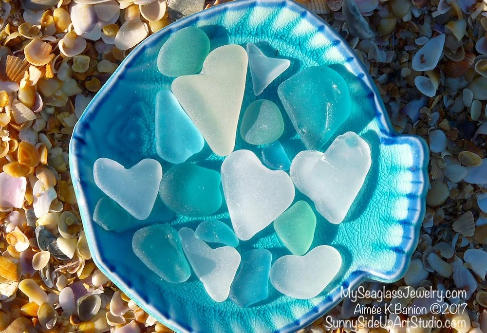 Sea glass hearts in shades of blue, green, and white on a blue shell-shaped dish, on a beach with small shells scattered around.