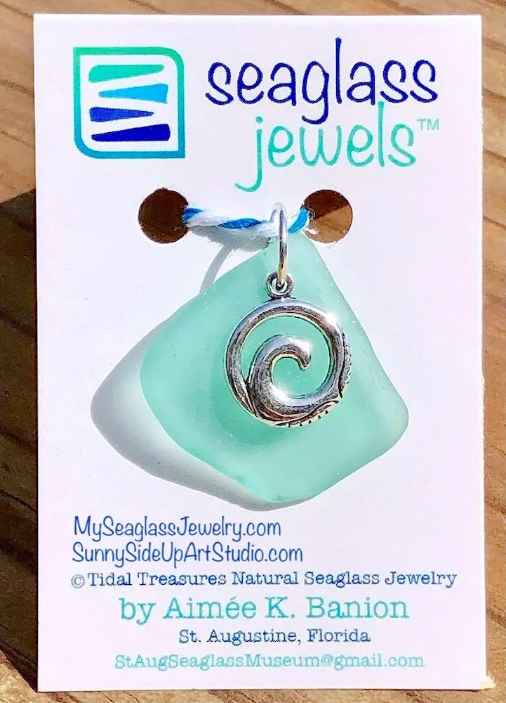 Seaglass jewelry piece with a silver spiral charm on a light green seaglass, displayed on a business card for Seaglass Jewels by Aimée K. Banion, featuring website links and contact information.