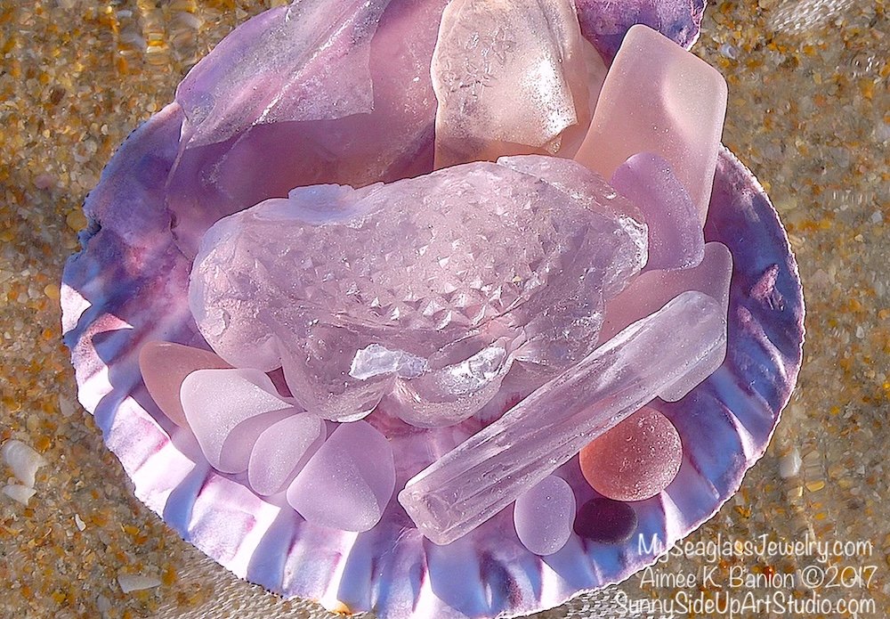Various lavender, purple and pink seaglass finds arranged on a purple iridescent shell on the beach.