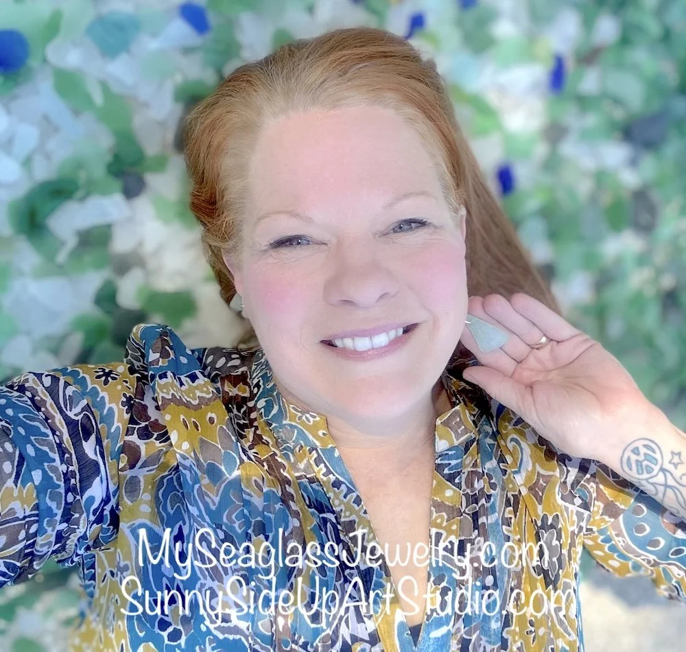 Close-up of seaglass jewelry artist Aimee Banion smiling, wearing a colorful patterned blouse, with a blurred seaglass background.