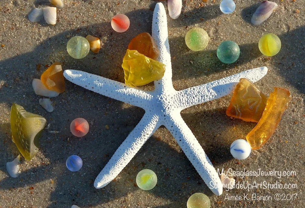 Starfish surrounded by colorful sea glass marbles and yellow sea glass on sand.
