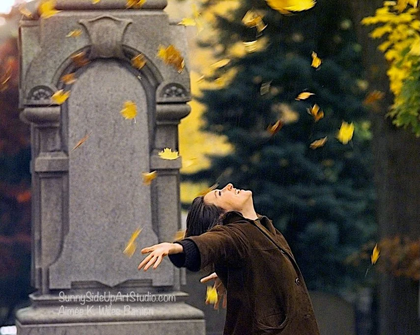 A woman in a brown jacket standing outdoors with her arms outstretched, head tilted back, and eyes closed as yellow leaves fall around her, photo by Aimee K. Wiles Banion while in Rochester, NY.