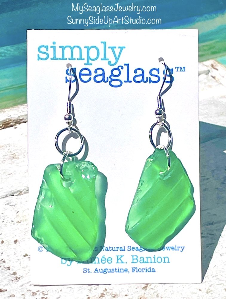 Green seaglass earrings with silver hooks hanging on a white display card featuring blue and turquoise text, outdoors with sand and water in the background.