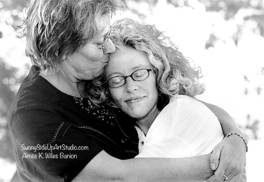 Two women, one older and one younger, sharing a warm hug and a kiss on the cheek outdoors, both with glasses and curly hair. Photographer Aimee Wiles Banion of Sunny Side Up Art + Photo makes candid photo portraits joyful, easy and fun. 