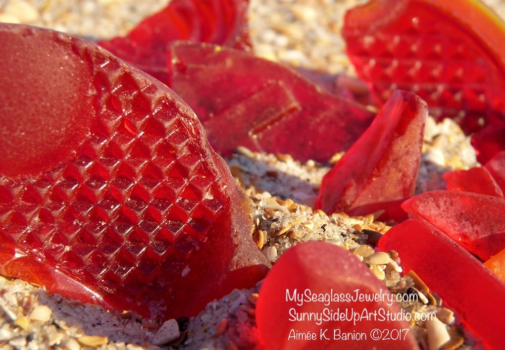 Close-up of red sea glass pieces on a sandy beach with small shells and pebbles.