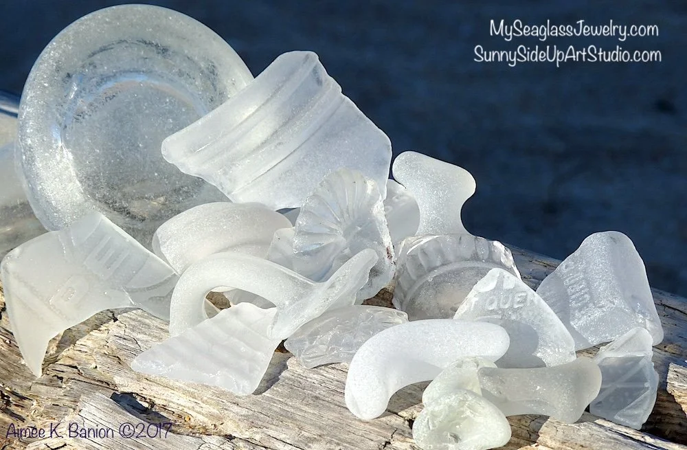 Collection of white frosty seaglass pieces on weathered wood with water in the background.