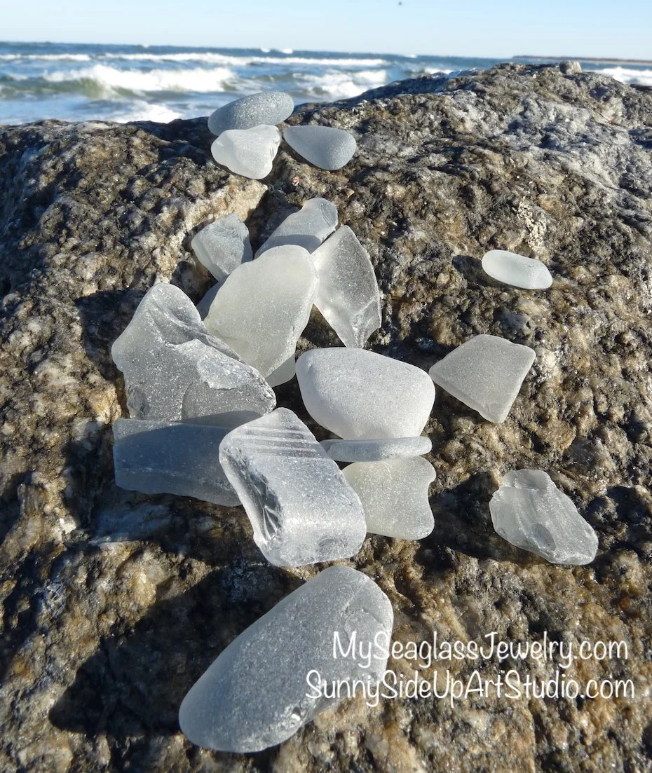 Collection of gray sea glass pieces on a rocky beach with ocean waves in the background.