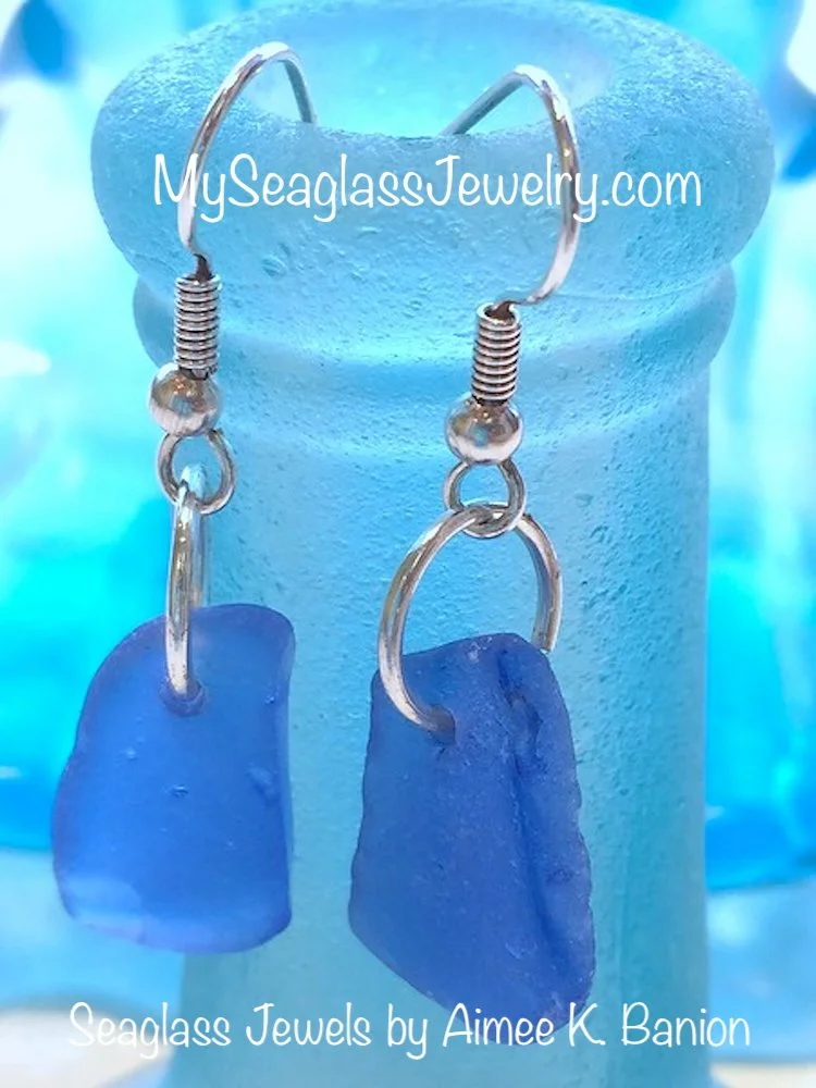 Blue sea glass earrings with metal hooks displayed on a blue stand.