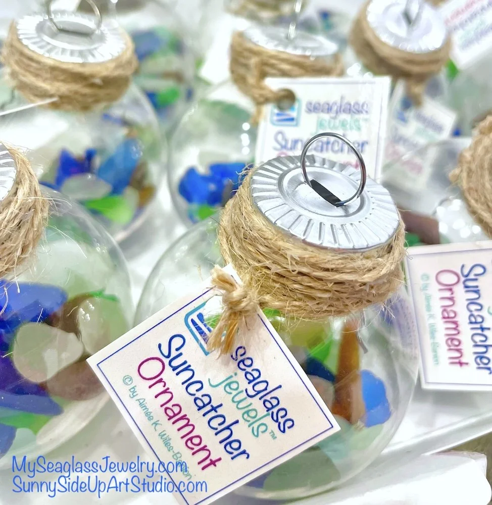 Clear glass ornaments filled with colorful sea glass, sealed with a metal lid wrapped in twine, and a tag attached that reads "Sea Glass Jewelry Suncatcher Ornament" from SunnySideUpArtStudio.com.