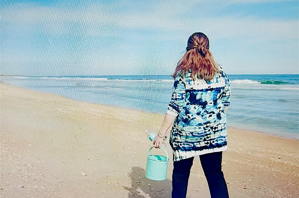 A woman with reddish-brown hair walking on a sandy beach holding a turquoise bucket, facing the ocean with waves, under a clear blue sky.