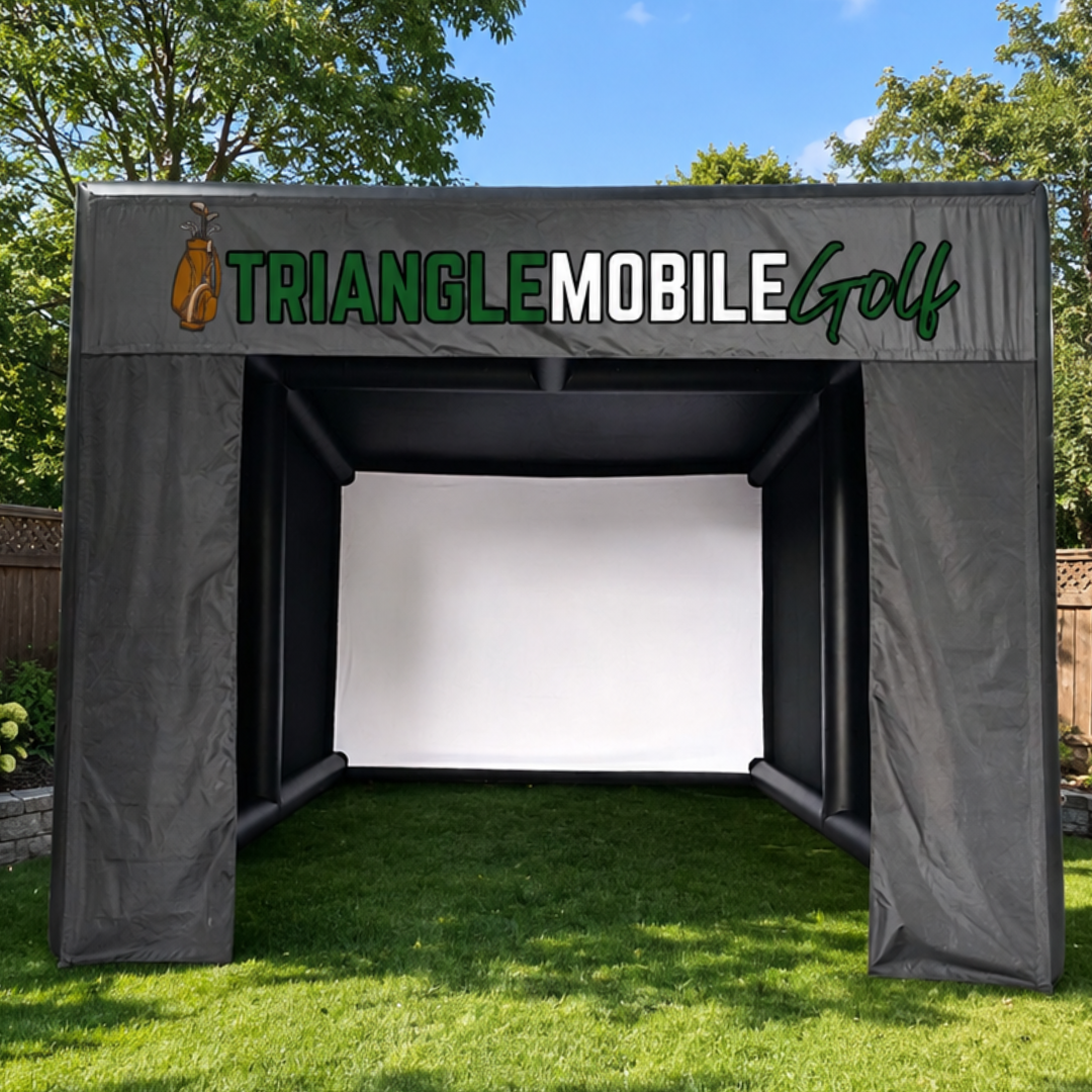 A golf driving range surrounded by a shelter with the logo 'Triangle Mobile Golf' on top, set in a grassy outdoor area with trees and a fence.