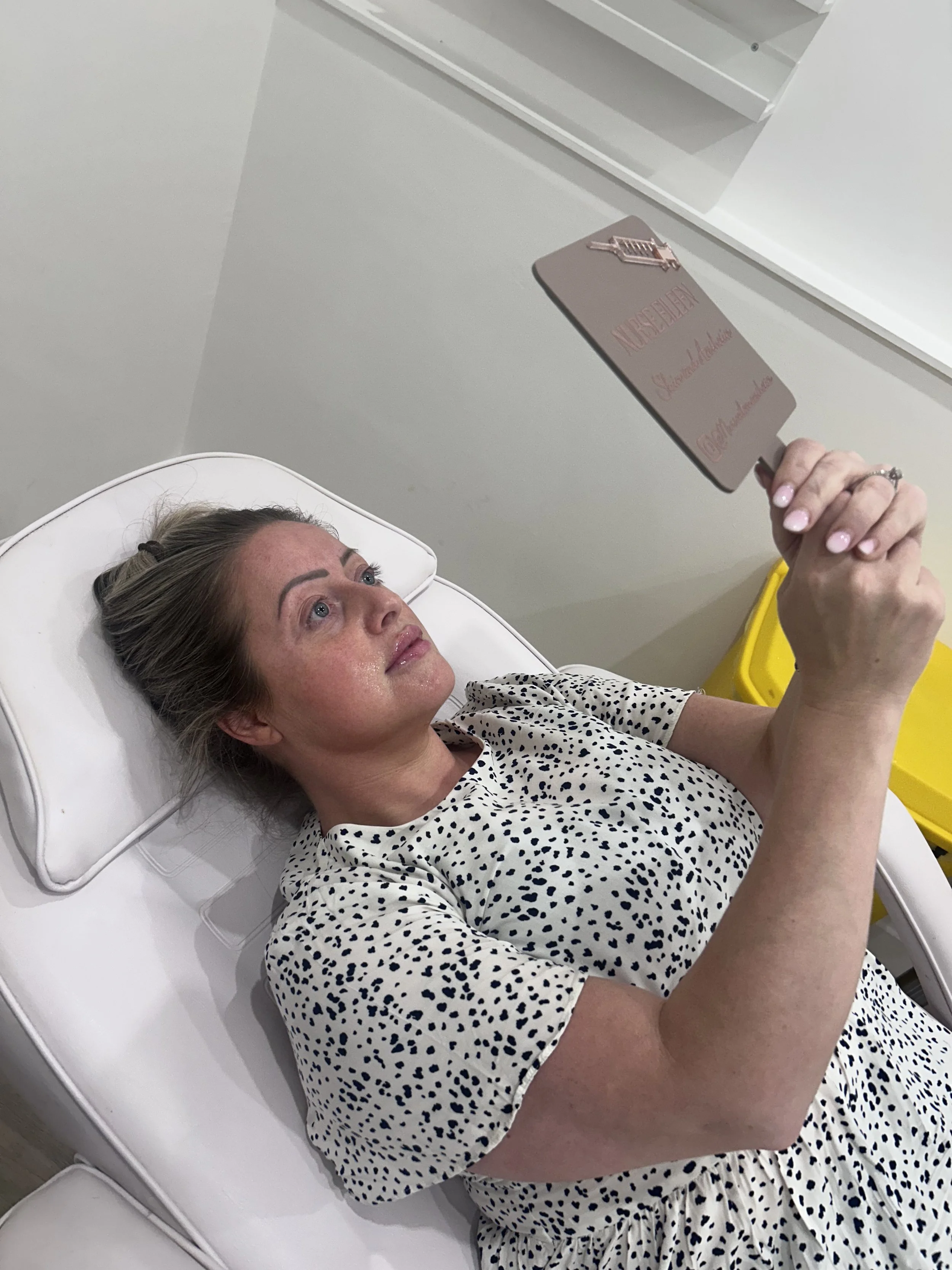 A woman with blonde hair, lying on a white medical bed, holding a brown sign with pink text that says 'Nurse Tech.' She is wearing a white dress with black polka dots and has a ring on her finger.