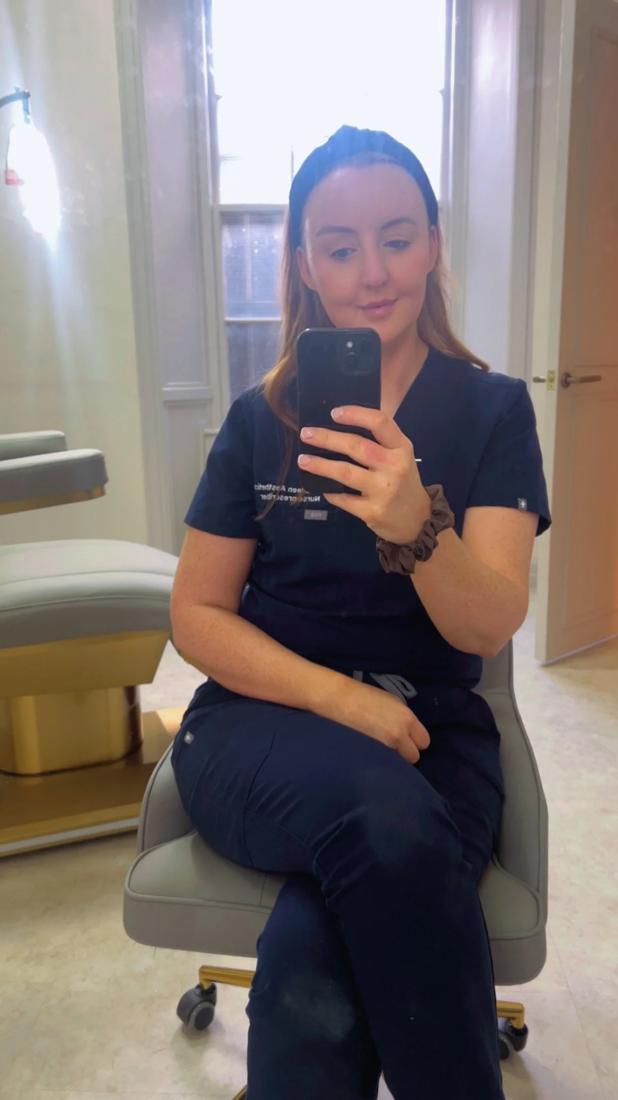 A woman taking a selfie with her phone in what appears to be a medical or dental office, sitting on a gray chair with beige flooring, and a medical equipment tray in the background.