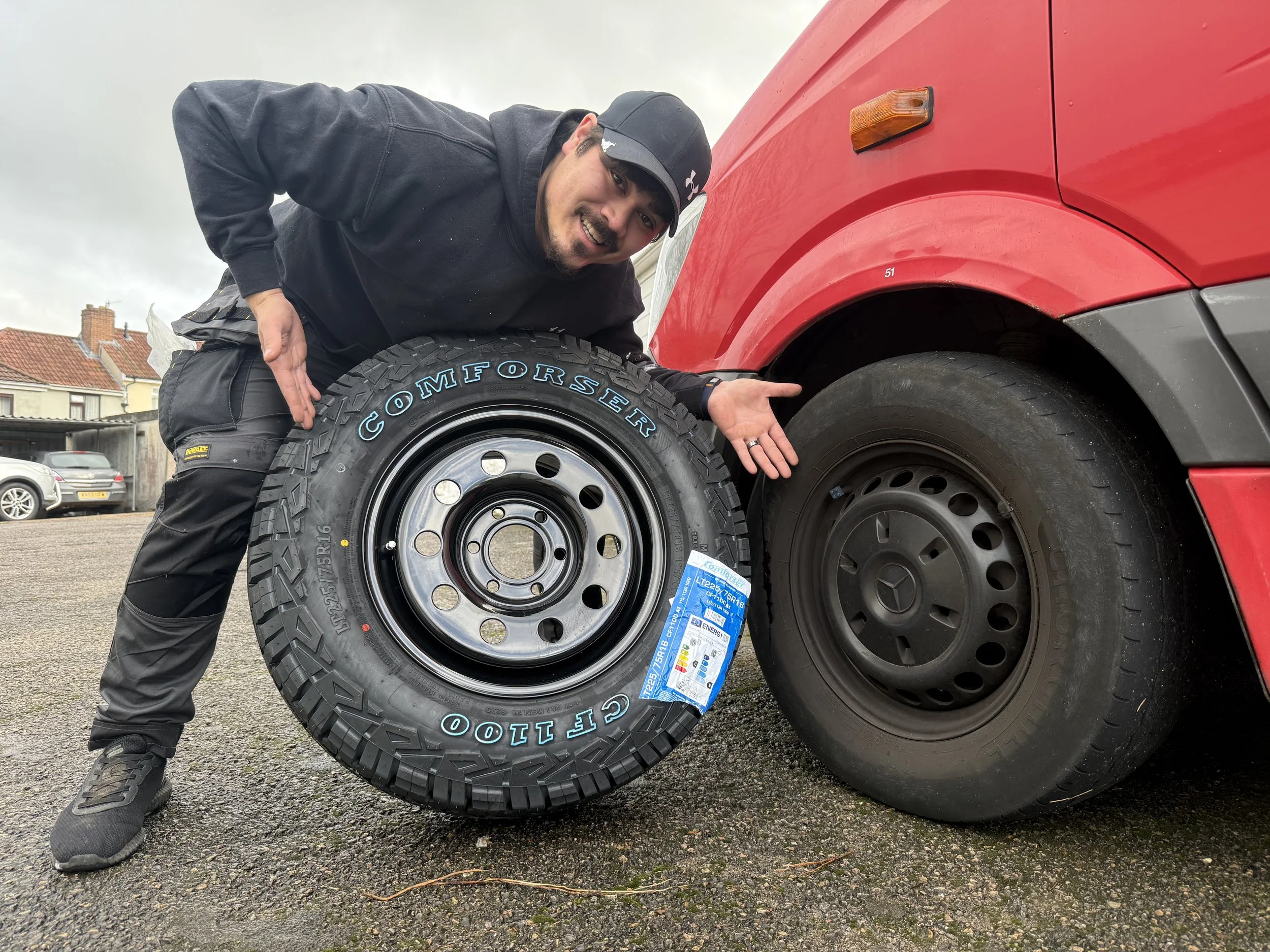 A man in black clothing and a cap crouches next to a red vehicle, holding a large tire with blue text on it. The background shows a parking lot with several cars and houses under a cloudy sky.