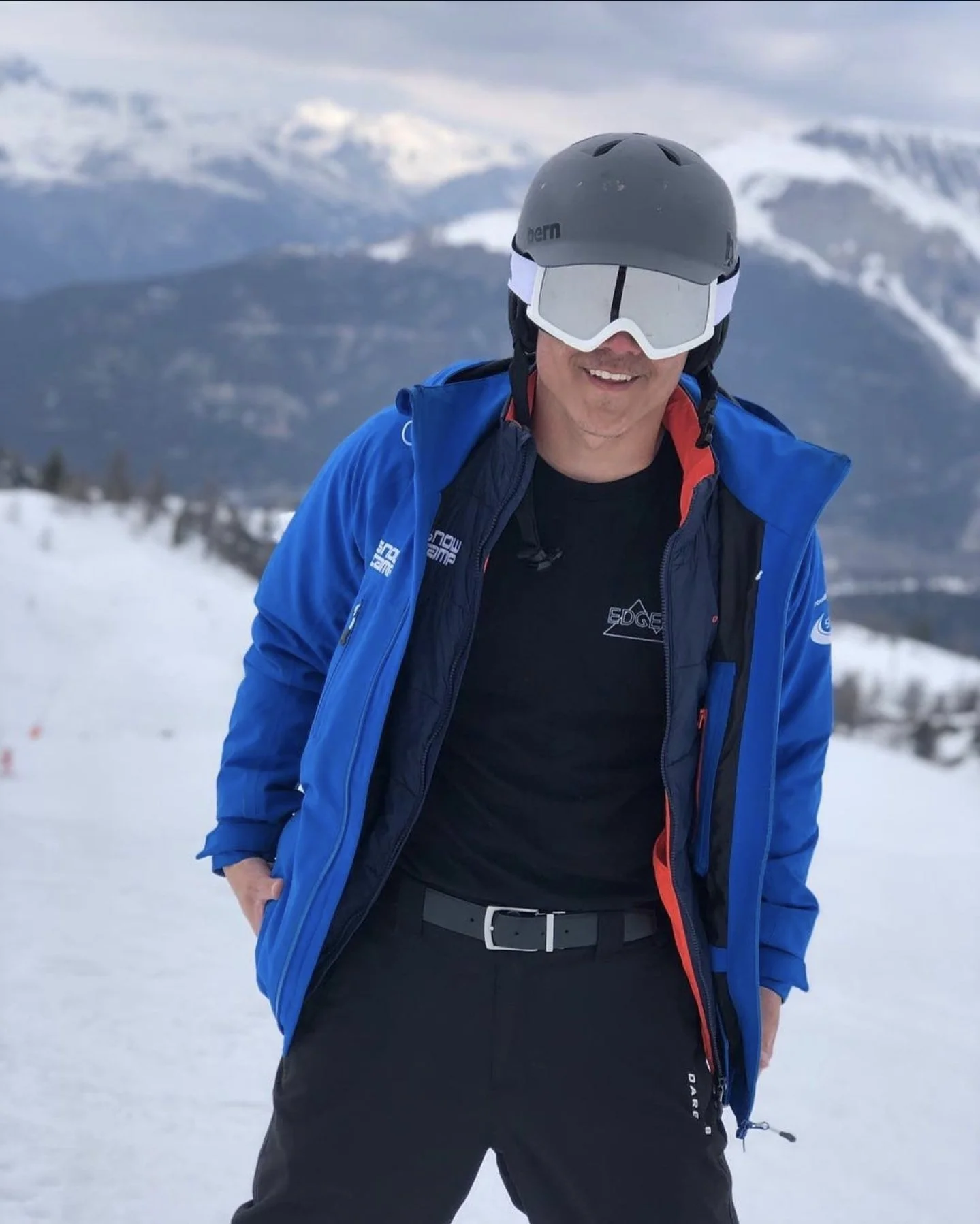 A man wearing a black helmet and white ski goggles. He is dressed in a blue jacket over a black shirt, standing in a snowy mountain landscape.