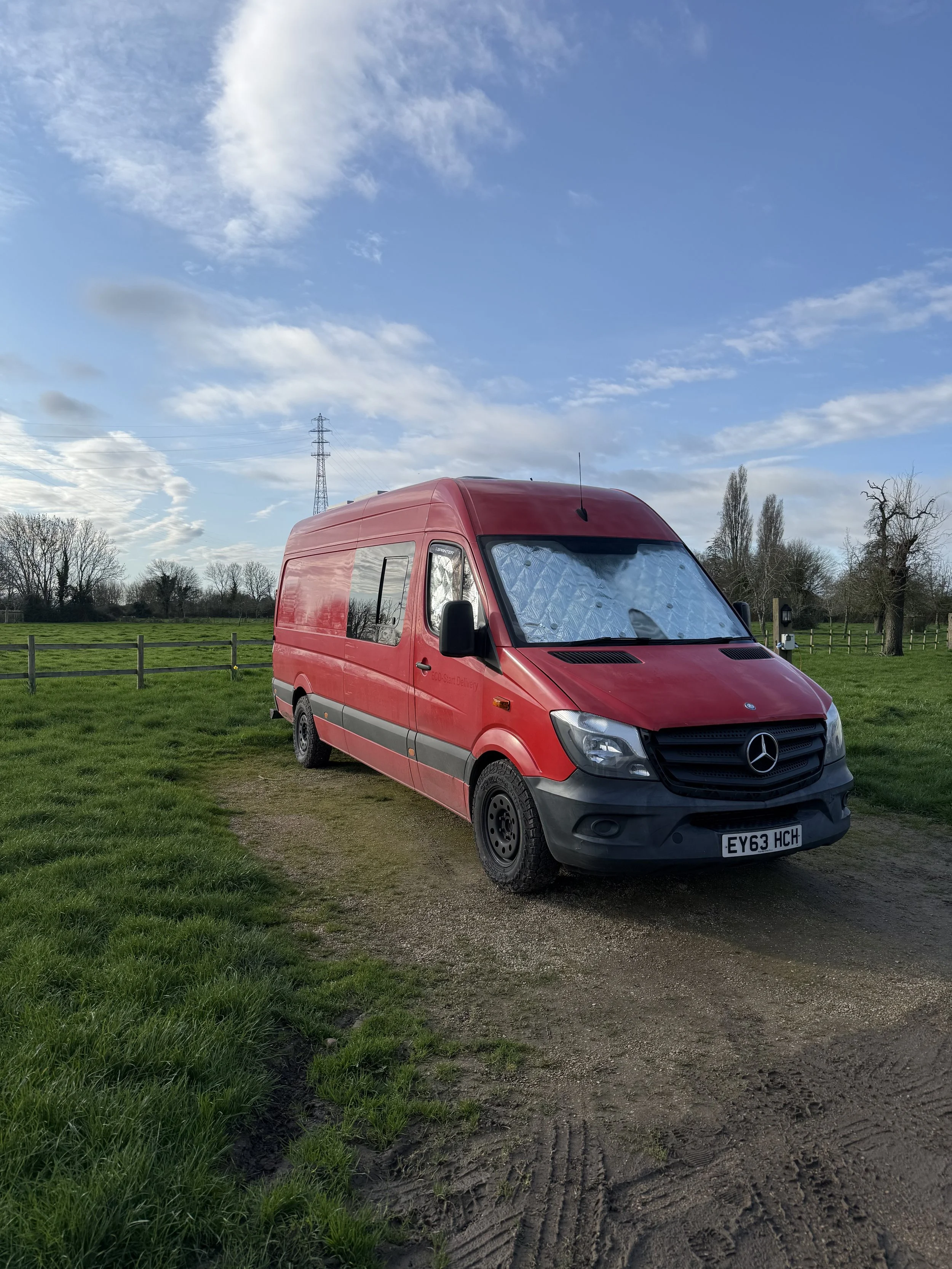 Red Mercedes-Benz van parked on a grassy field with a dirt path, trees, a wooden fence, and a cloudy sky in the background.
