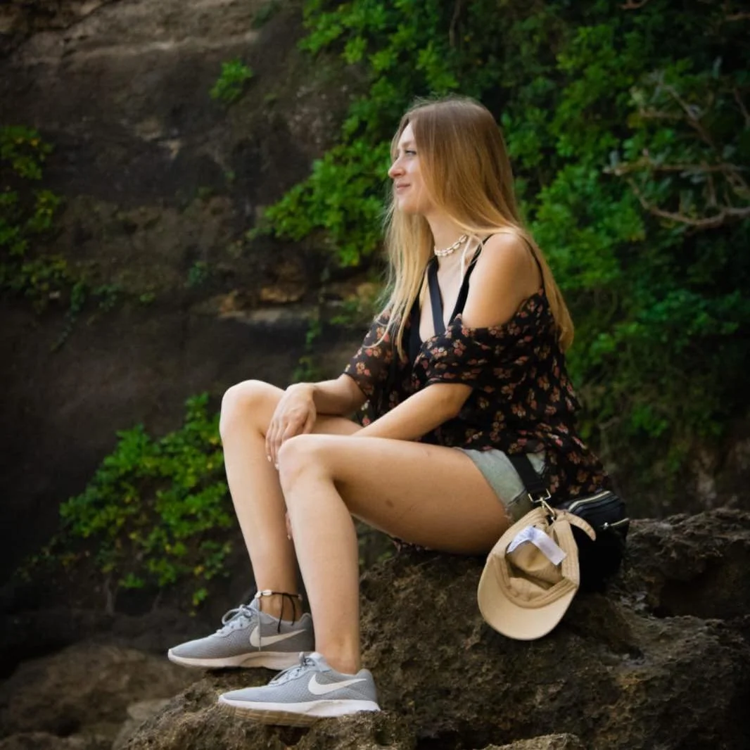 A young woman sitting on a rock outdoors, wearing a black floral top, denim shorts, gray Nike sneakers, and a necklace, with a beige hat and black bag beside her, surrounded by green bushes.