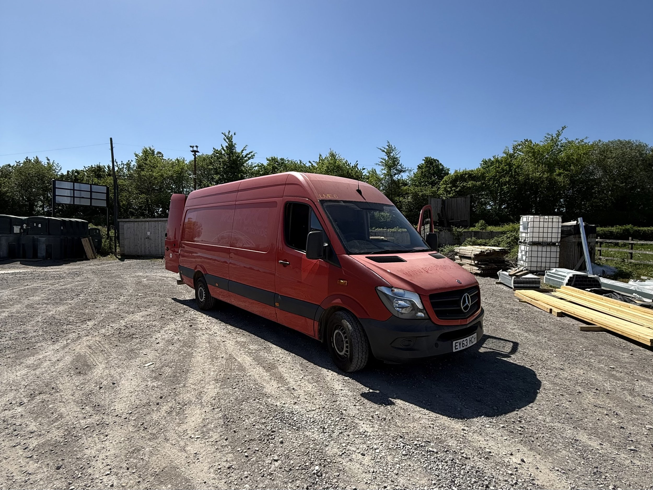A red Mercedes-Benz van parked on a gravel lot with trees, construction materials, and a wooden fence in the background under a clear blue sky.