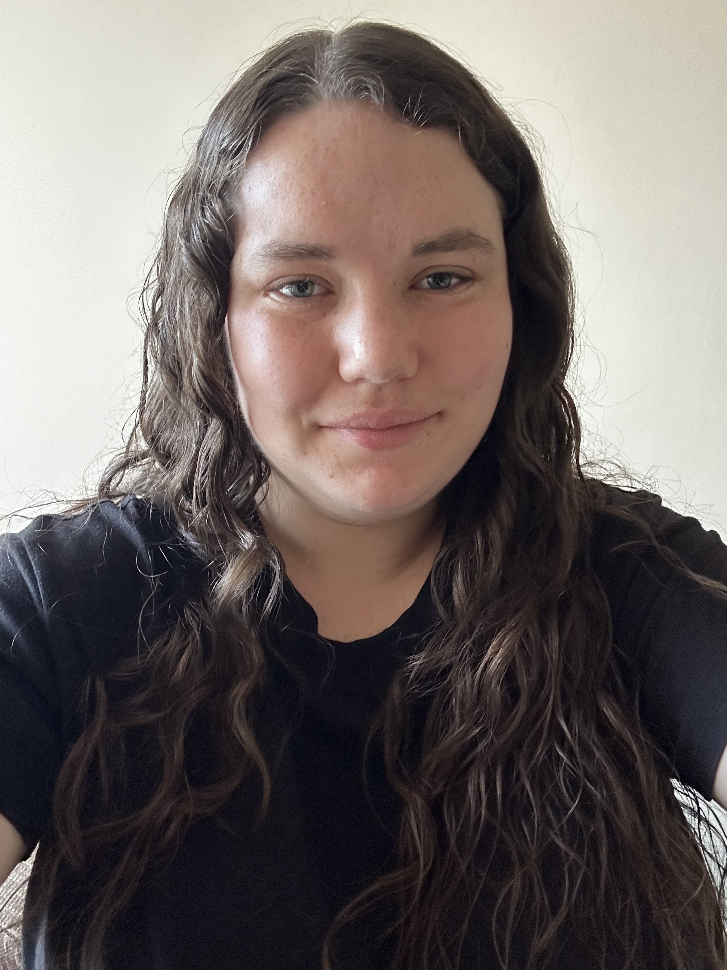 A woman with long, wavy brown hair, light skin, and blue eyes smiling softly, wearing a black shirt against a plain light-colored wall.