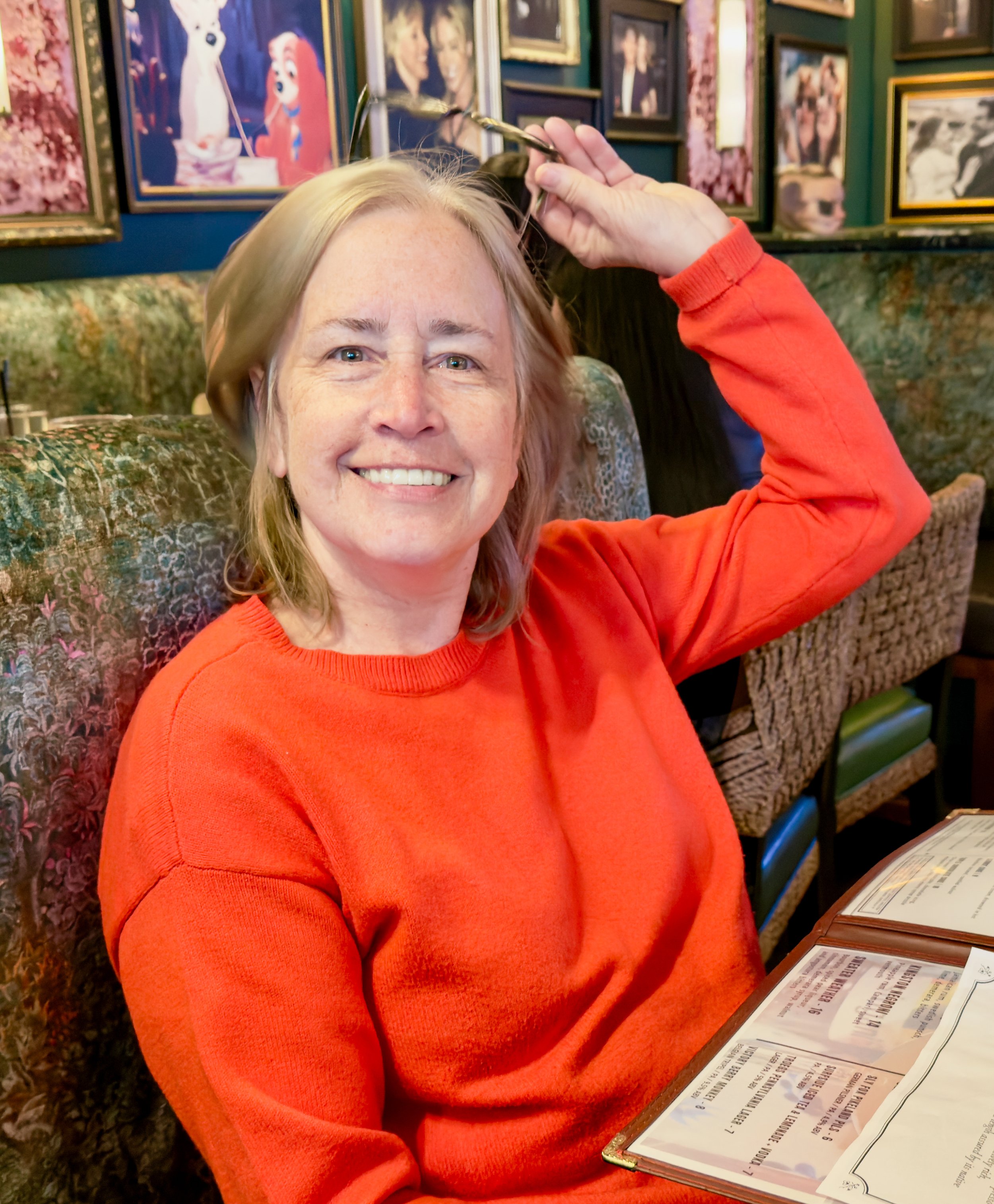 A photograph of artist and author, Karen Serago, smiling with blonde hair and a red sweater sitting at a restaurant table, holding glasses above her head. The background features framed pictures of animals and people on a green wall.