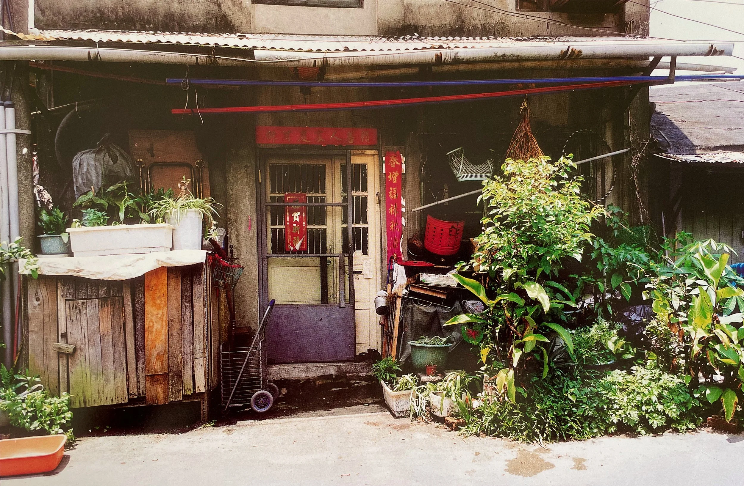 A cluttered entrance of an old house with potted plants, gardening tools, and red decorative banners with Chinese characters, surrounded by greenery and a concrete pathway, photographed by artist, Karen Serago.