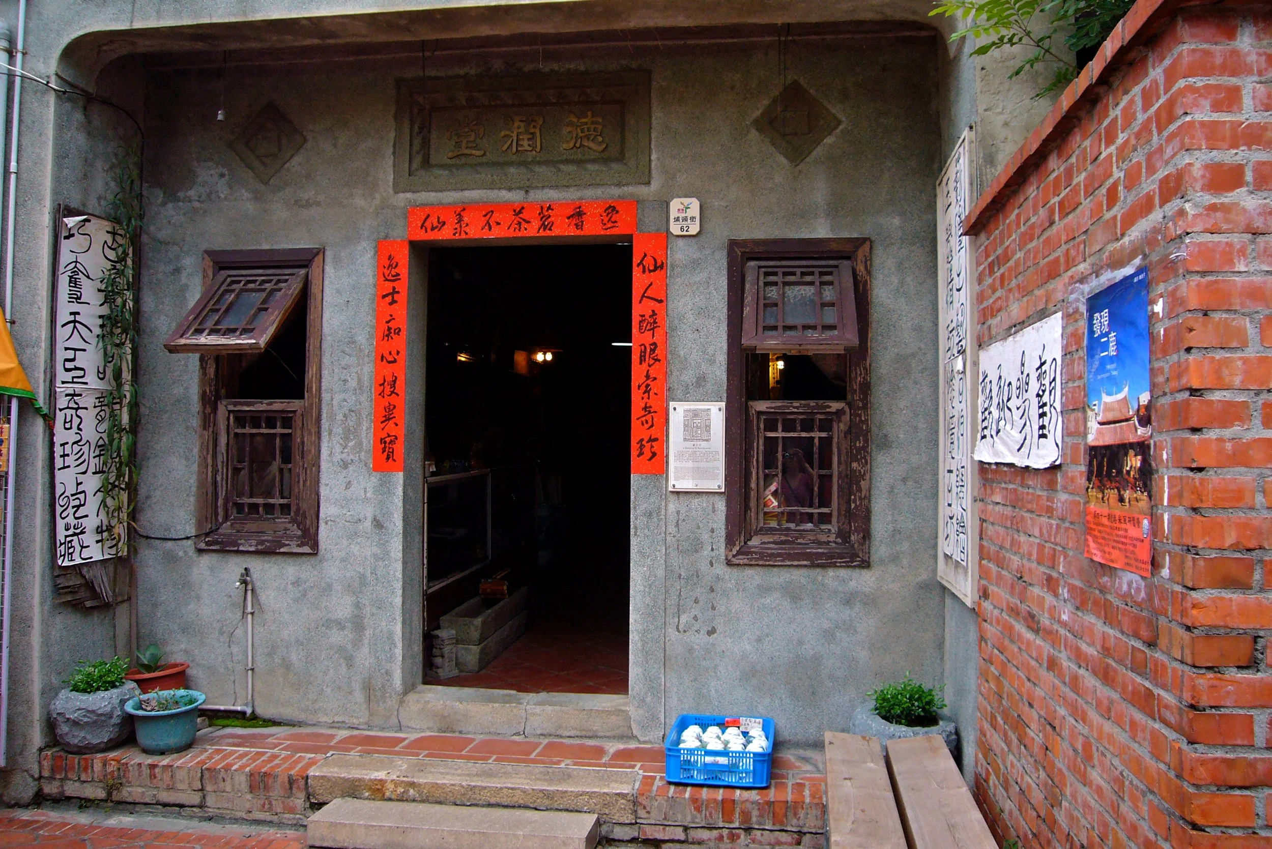 View of a building entrance with concrete and brick walls, open wooden window shutters, potted plants, and Asian calligraphy signs. Inside, warm lighting is visible, photographed by artist, Karen Serago.