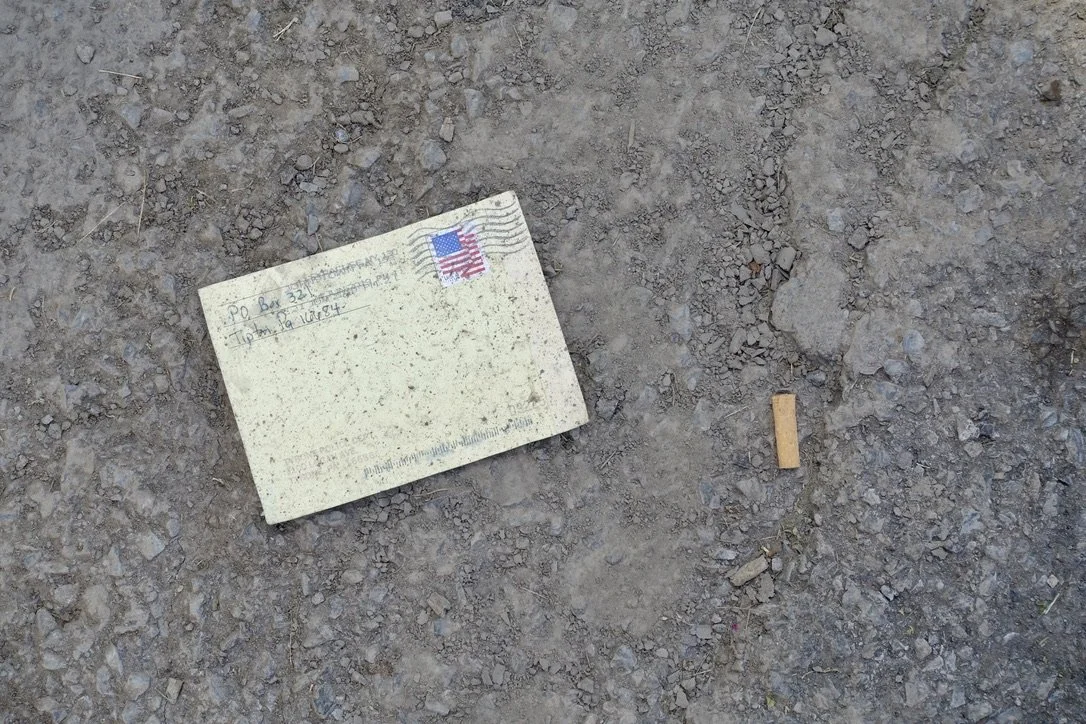 A discarded cigarette pack and a small cigarette on dirt ground, photographed by artist, Karen Serago.