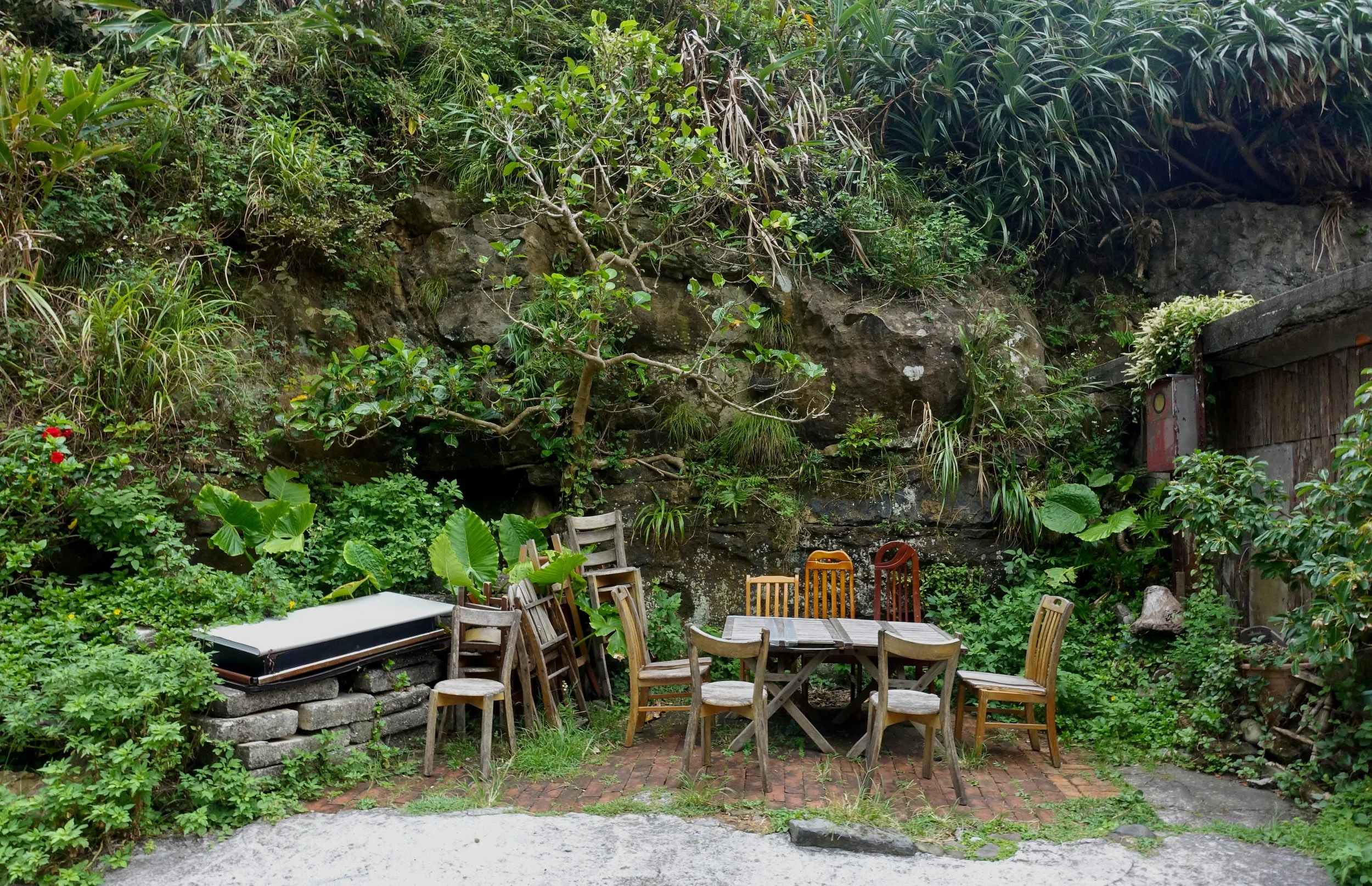 An outdoor seating area with wooden chairs and tables, surrounded by lush green plants, trees, and rocks, creating a natural and cozy garden setting, photographed by artist, Karen Serago.