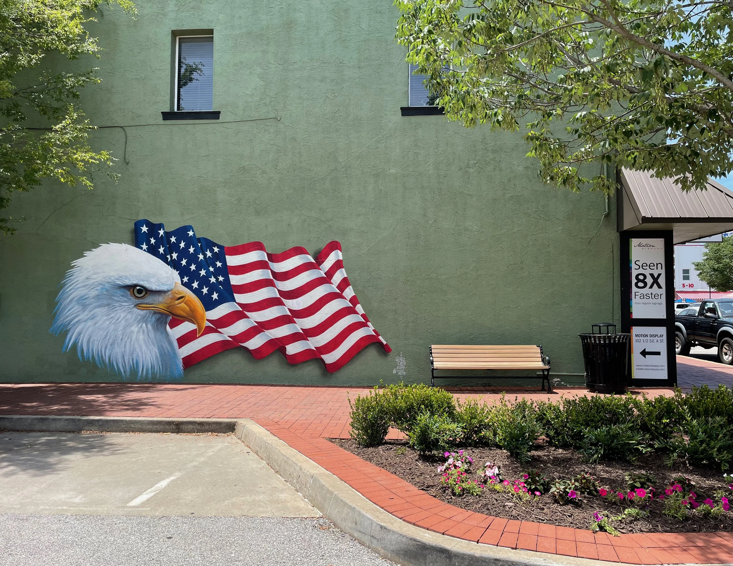 Part of a mural of a bald eagle with an American flag in its beak in front of a green building with two windows, trees, a bench, and a parking lot photographed by artist, Karen Serago.