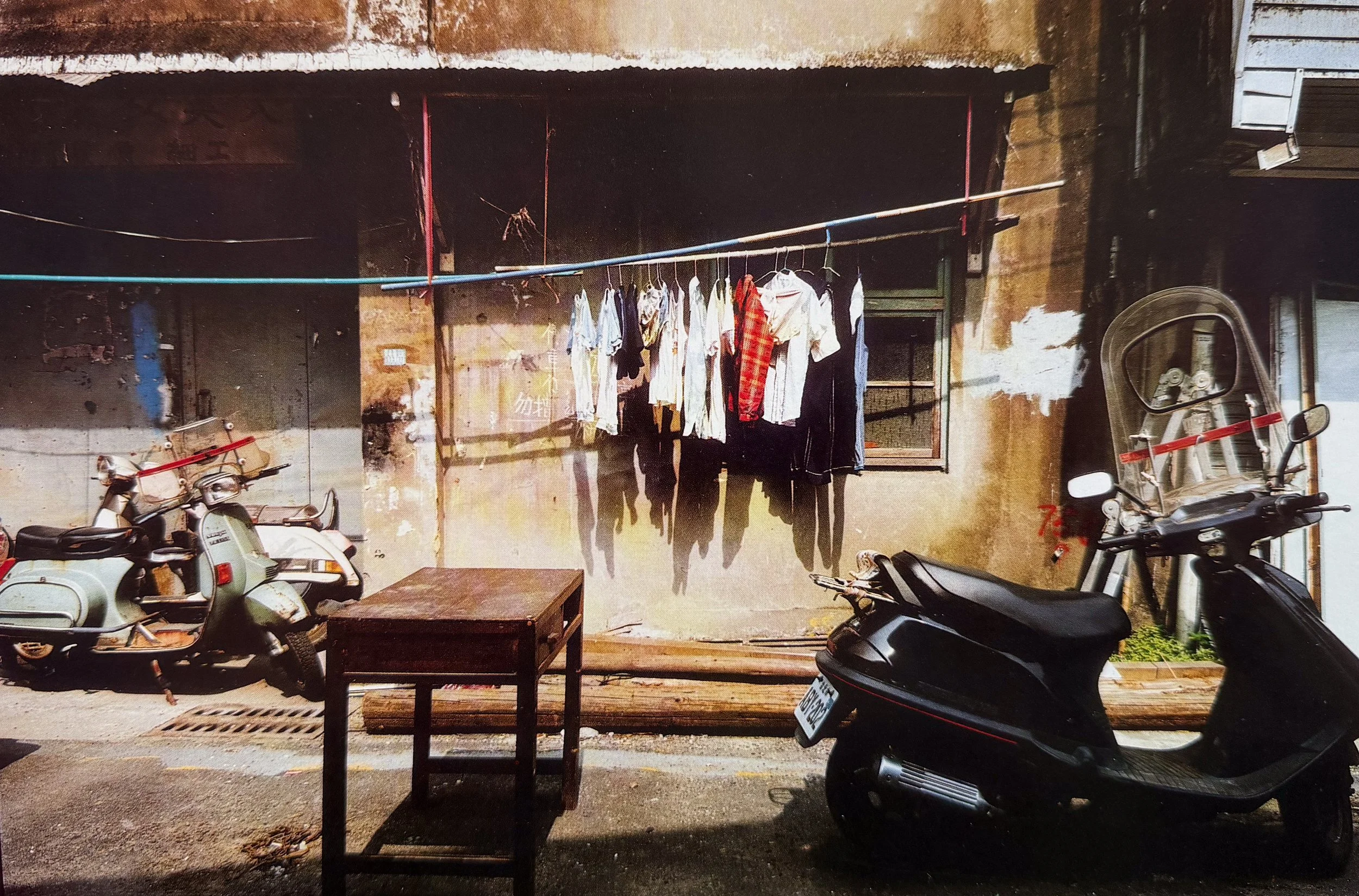 Clothes hanging on a clothesline outside a building, with scooters parked underneath and a wooden table in front, photographed by visual artist, Karen Serago.
