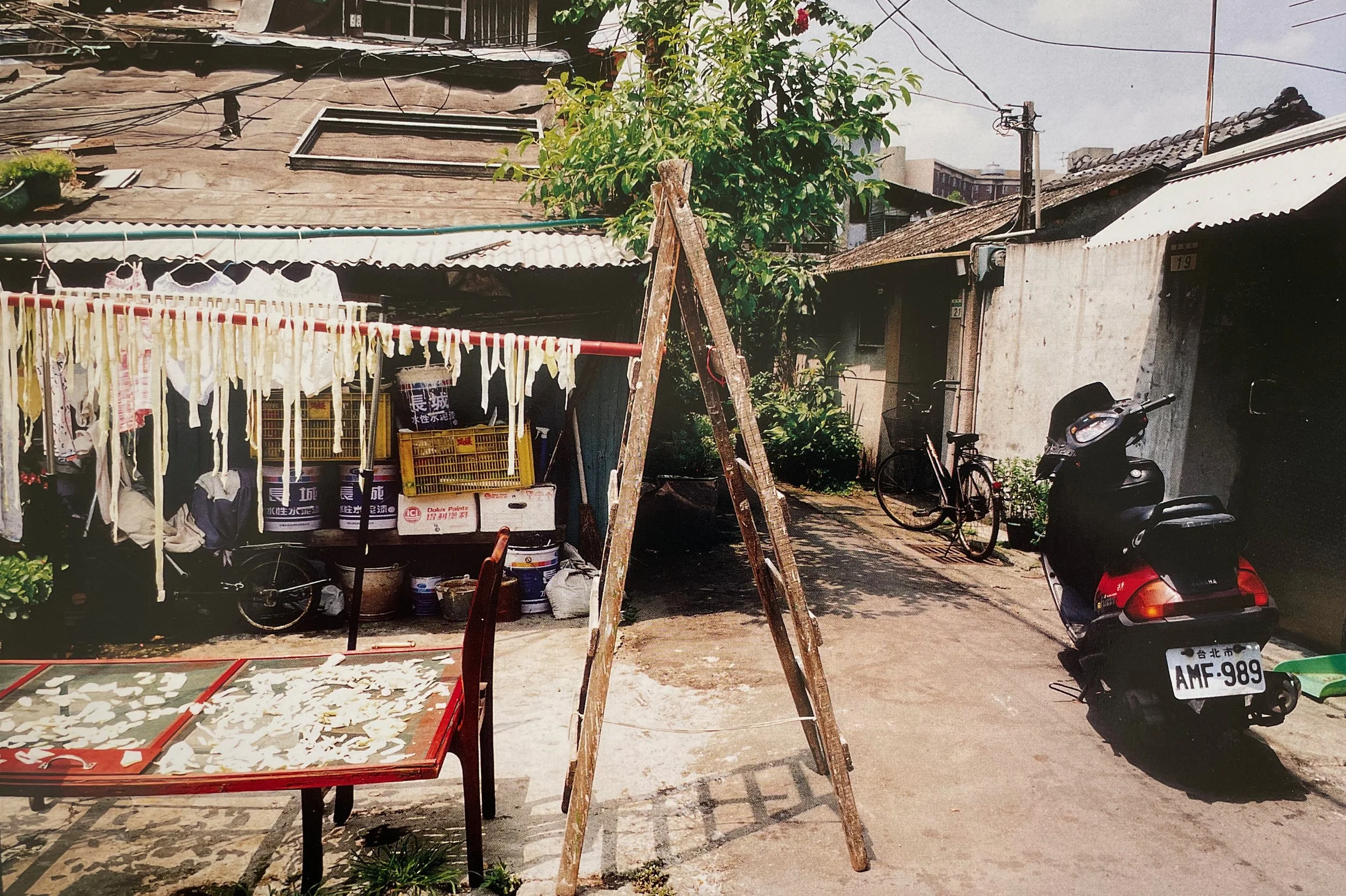 Street scene with laundry hanging on a clothesline, bicycles, motorbikes, and old buildings with tiled roofs, trees, and power lines, photographed by artist, Karen Serago.