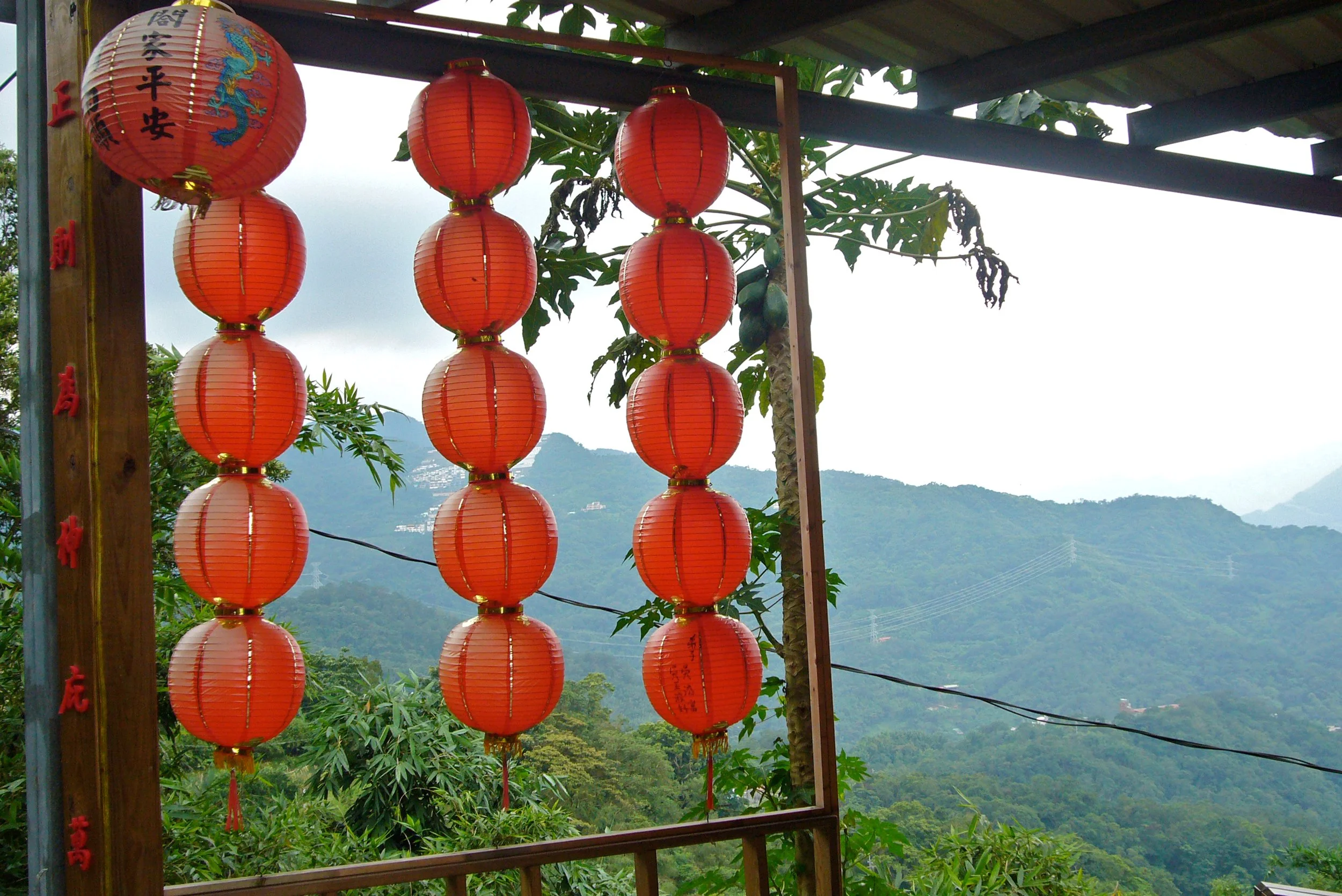 Multiple red Chinese lanterns hanging outdoors against a mountain landscape backdrop, photographed by artist, Karen Serago.
