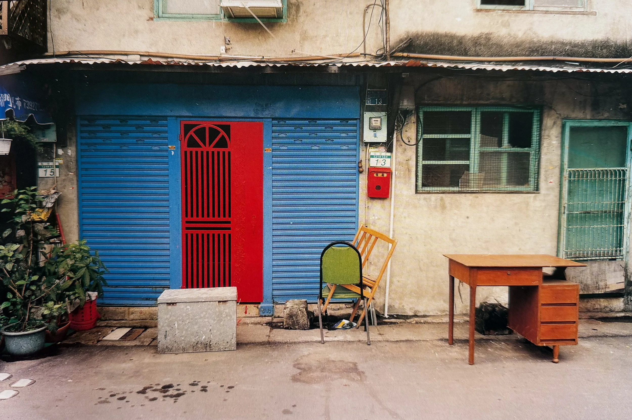 An outdoor scene with a blue metal rolling door that has a red gate design. To the right, there is a window with a green metal grid and a small white box mounted on the wall. In front, there are two chairs, one green and one orange, along with a wooden table. Potted plants are visible on the left side, and miscellaneous objects are placed along the ground and around the scene.