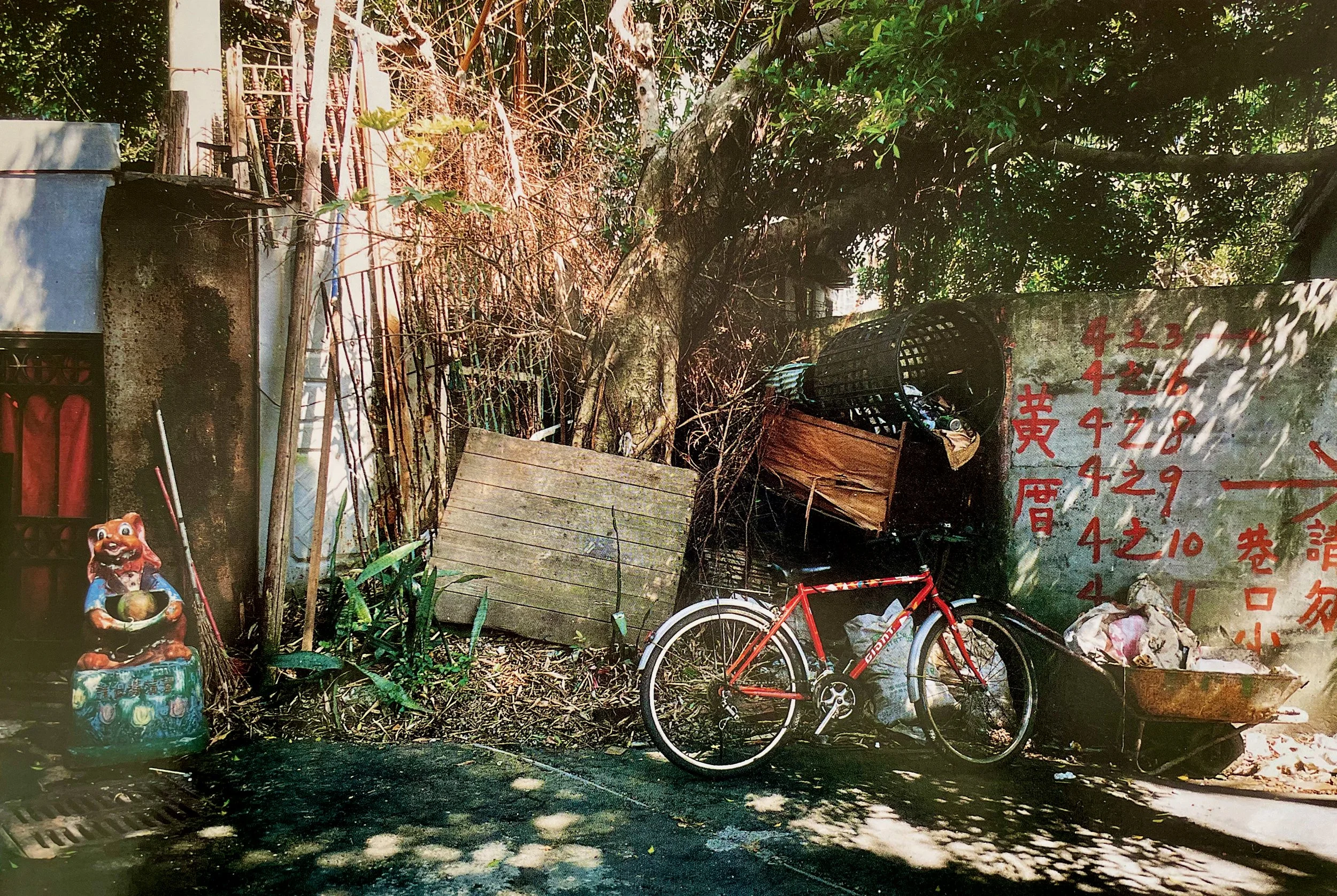 An outdoor scene with a bicycle leaning against a wall with red Chinese characters, a small decorative statue of a dog, and various objects like a wooden board and metal grates, trees and plants. Photographed by visual artist, Karen Serago.