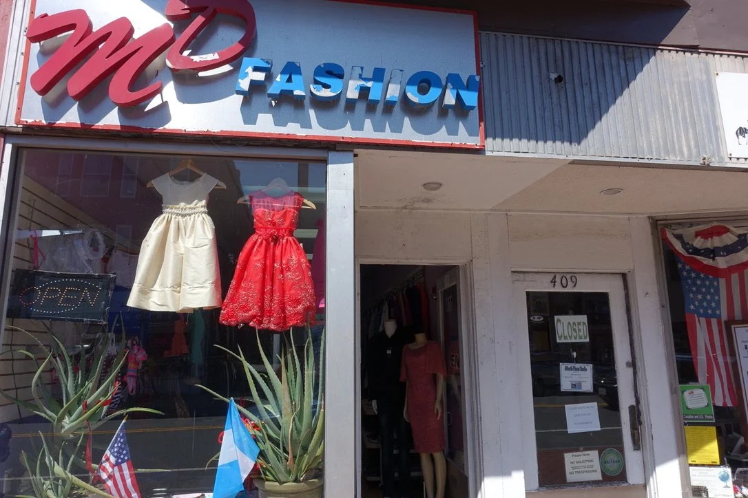 The storefront of M&D Fashion with mannequins inside displaying dresses, one white and one red, and a sign indicating the shop is closed, photographed by artist, Karen Serago.