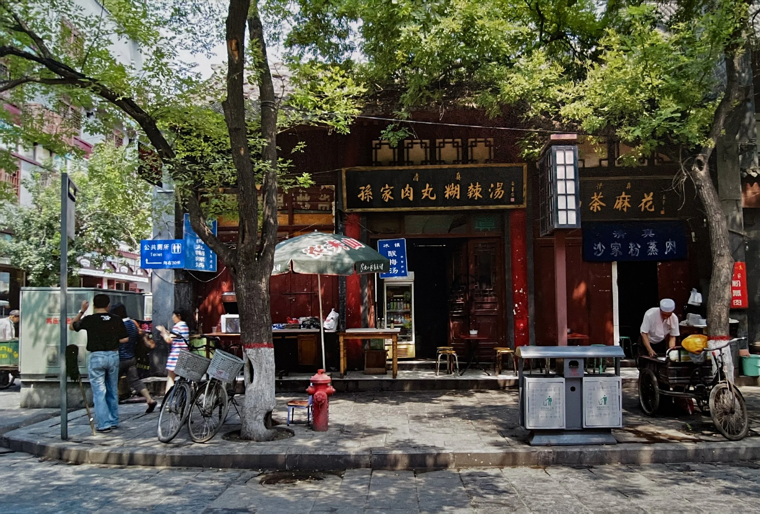 Outdoor scene with people near a traditional Taiwanese building, trees, bicycles, and street signs in Chinese and English. Photographed by visual artist, Karen Serago.