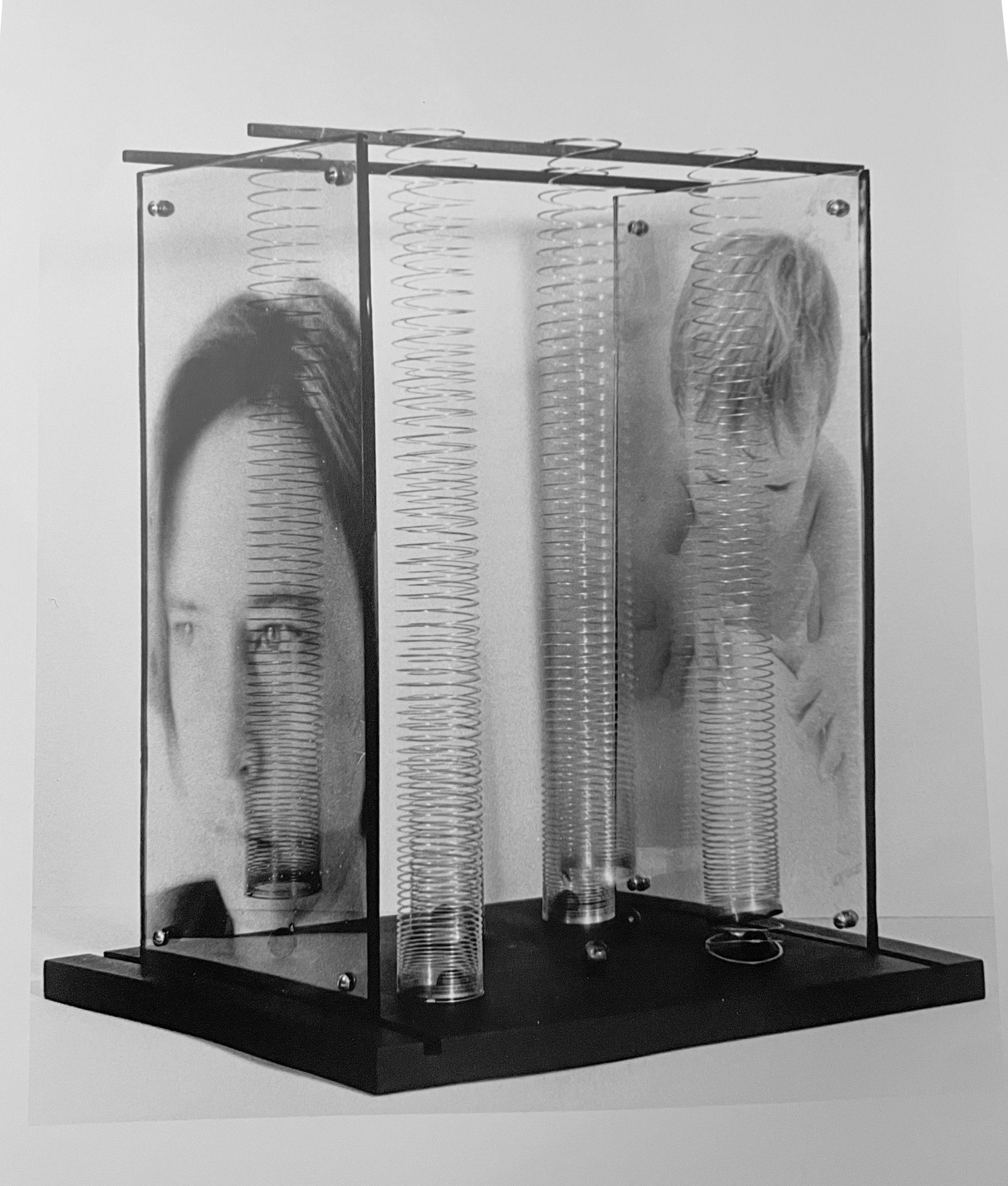 A scientific apparatus featuring three transparent vertical chambers with spiral metal coils inside, mounted on a black base with metal supports and connectors, by artist, Karen Serago.