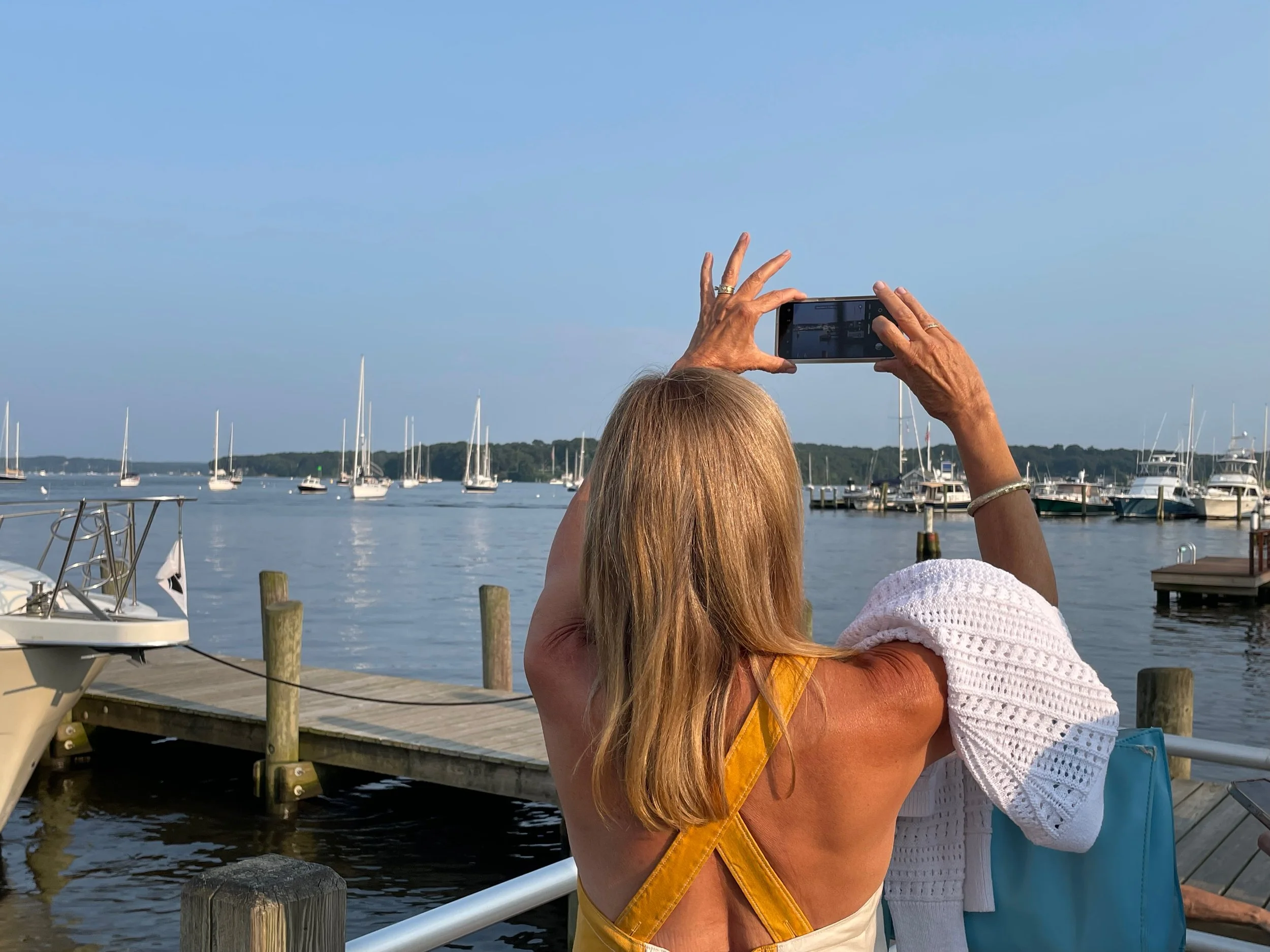 A person with long blonde hair is taking a photo of sailboats on a marina with a smartphone, seen from behind. The person is wearing a sleeveless top and a yellow bag strap, standing on a dock near water, photographed by artist, Karen Serago.