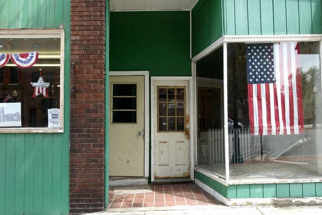 A storefront with green siding, brick column, and a large American flag in the window. There are two weathered white doors and a window display with patriotic decorations, photographed by artist, Karen Serago.