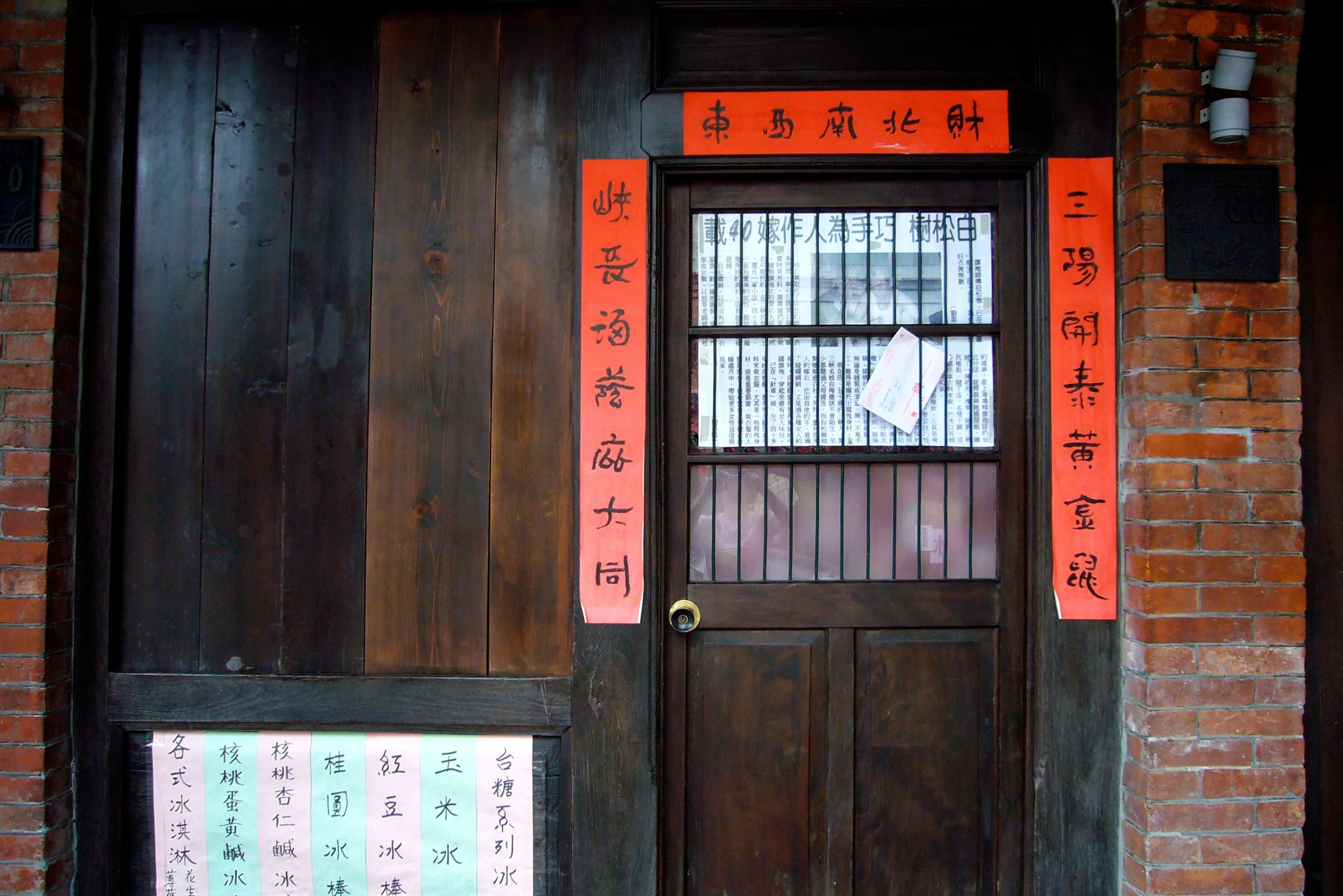 A wooden door framed by red brick walls, with red vertical and horizontal banners with Chinese characters on either side and above the door. The door has a small barred window with papers and signs attached, photographed by artist, Karen Serago.